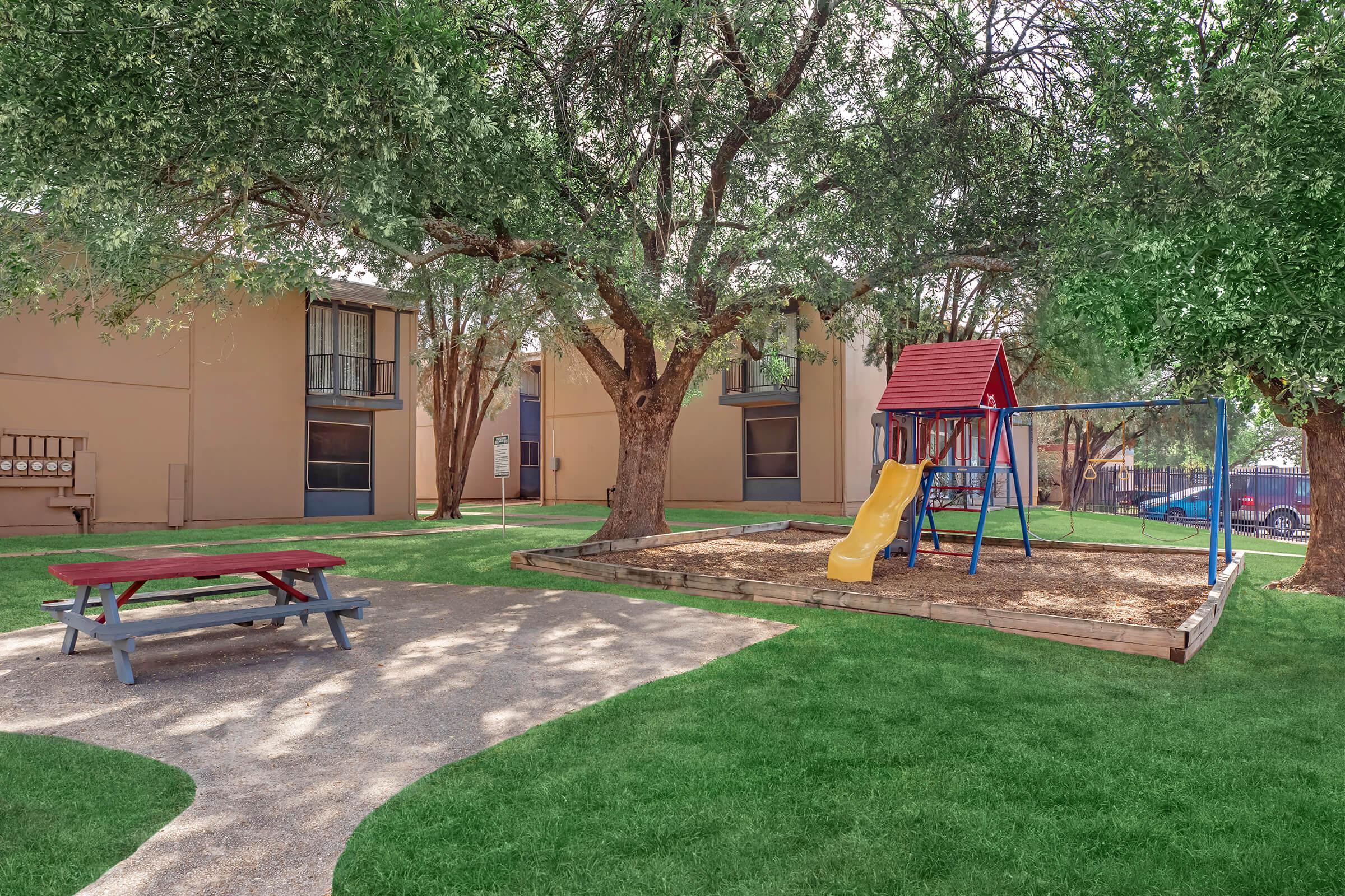 A playground featuring a yellow slide and swing set, surrounded by grass and trees. Nearby is a picnic table on a pathway. The scene includes two buildings in the background, creating a welcoming outdoor area for children and families.