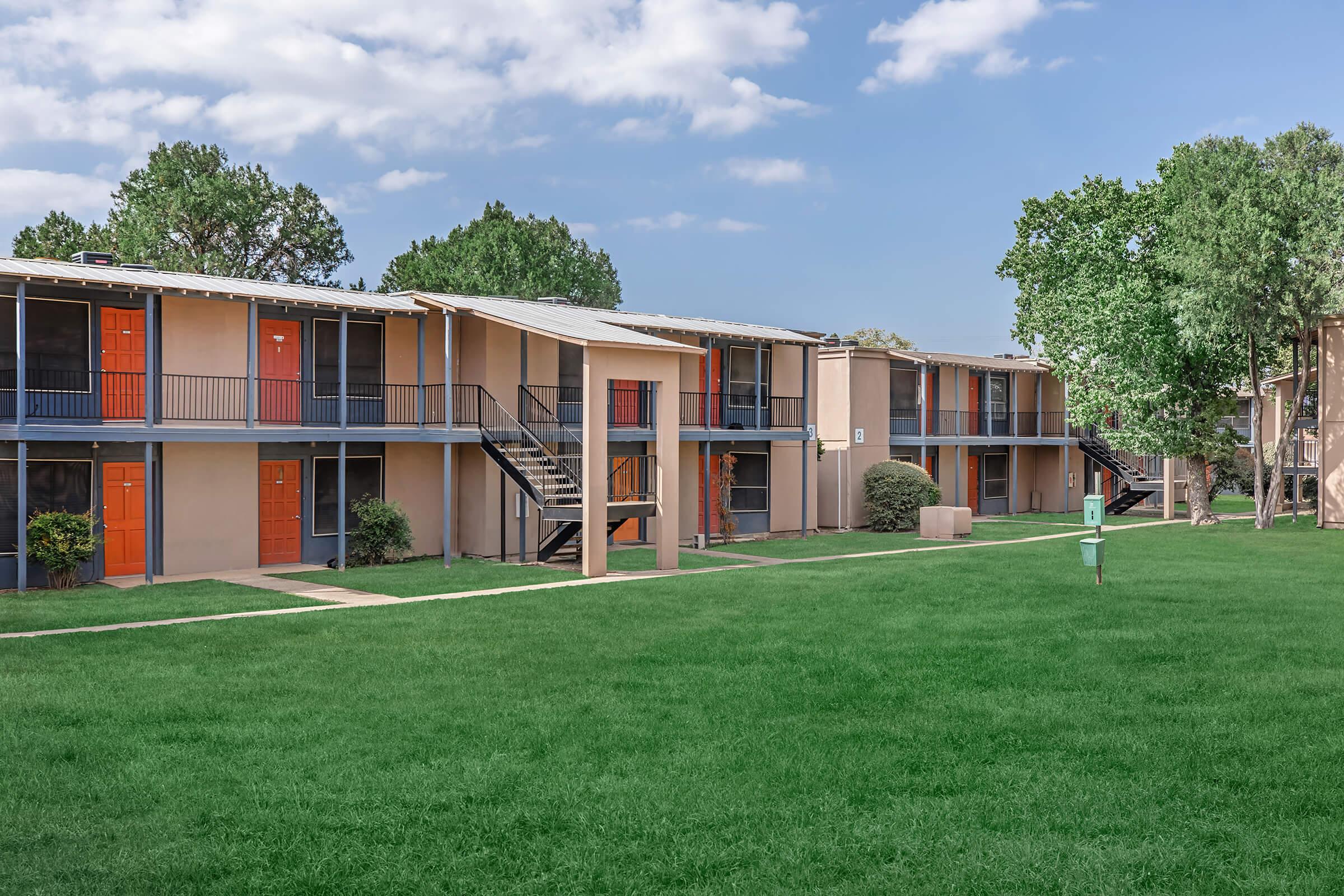 A well-maintained apartment complex featuring two-story buildings with orange doors. The grassy courtyard is lined with neatly trimmed shrubs and trees, and a staircase leads to the upper units. The sky is clear with a few clouds, creating a bright and inviting atmosphere.