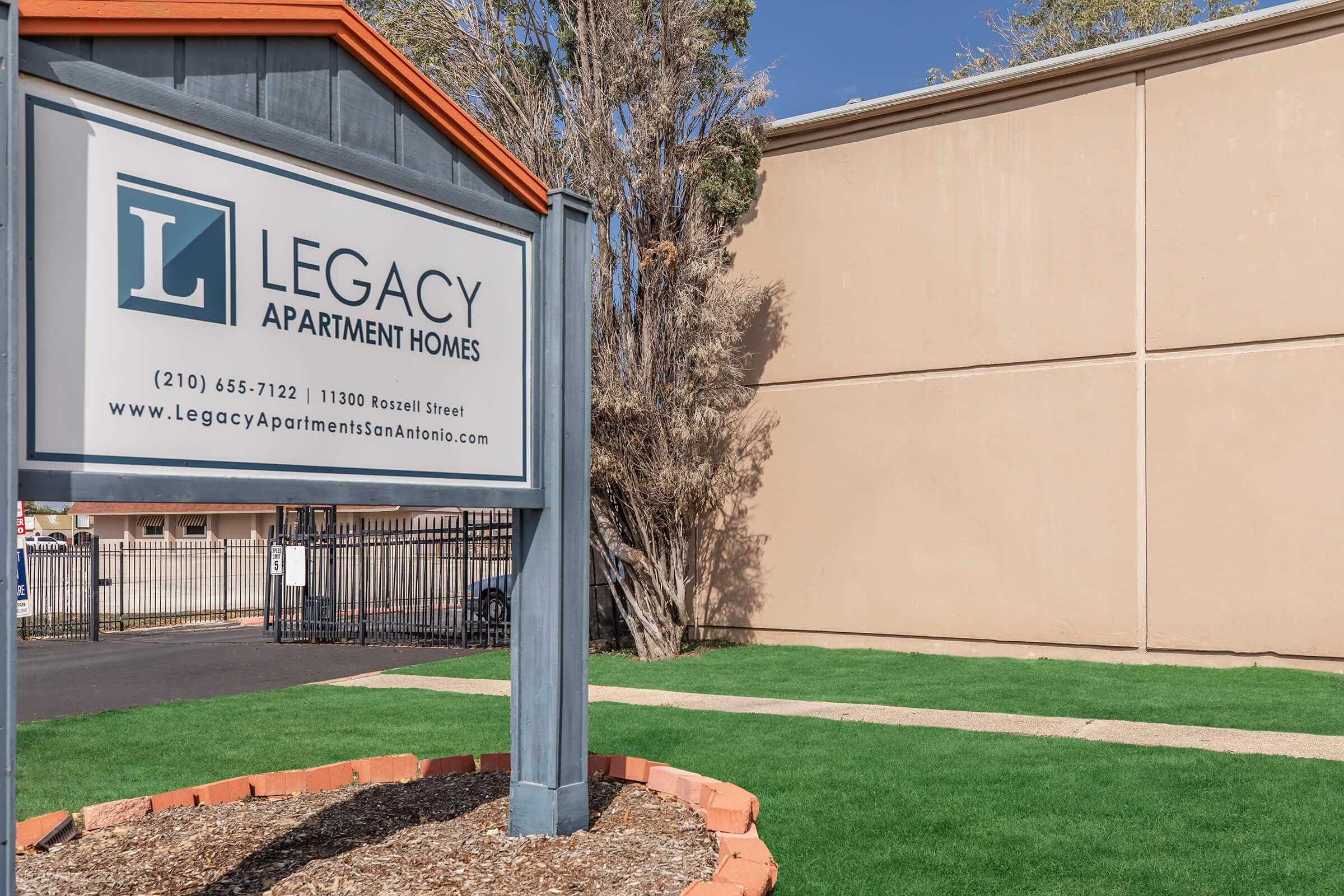 Sign for Legacy Apartment Homes at 11300 Roszell Street, featuring a large "L" logo. The sign is situated in front of a well-maintained green lawn, with a fence visible in the background. The image conveys a welcoming atmosphere for potential residents.