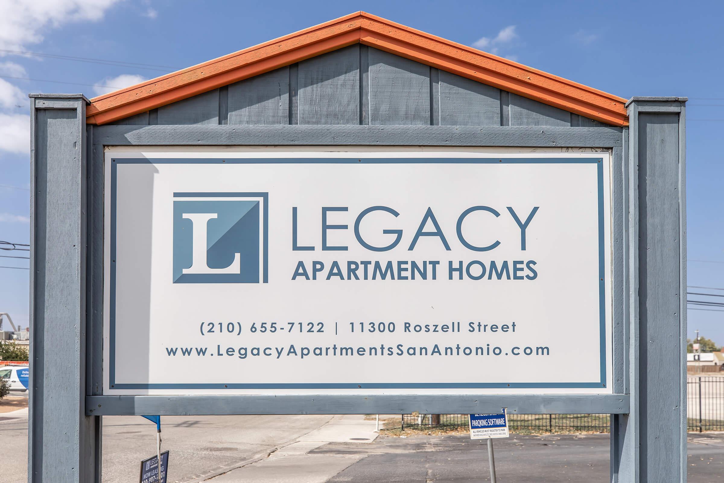 Sign for Legacy Apartment Homes, displaying the phone number (210) 655-7122, the address 11300 Roszell Street, and the website LegacyApartmentsSanAntonio.com. The sign features a blue and white design with the letter "L" prominently displayed.