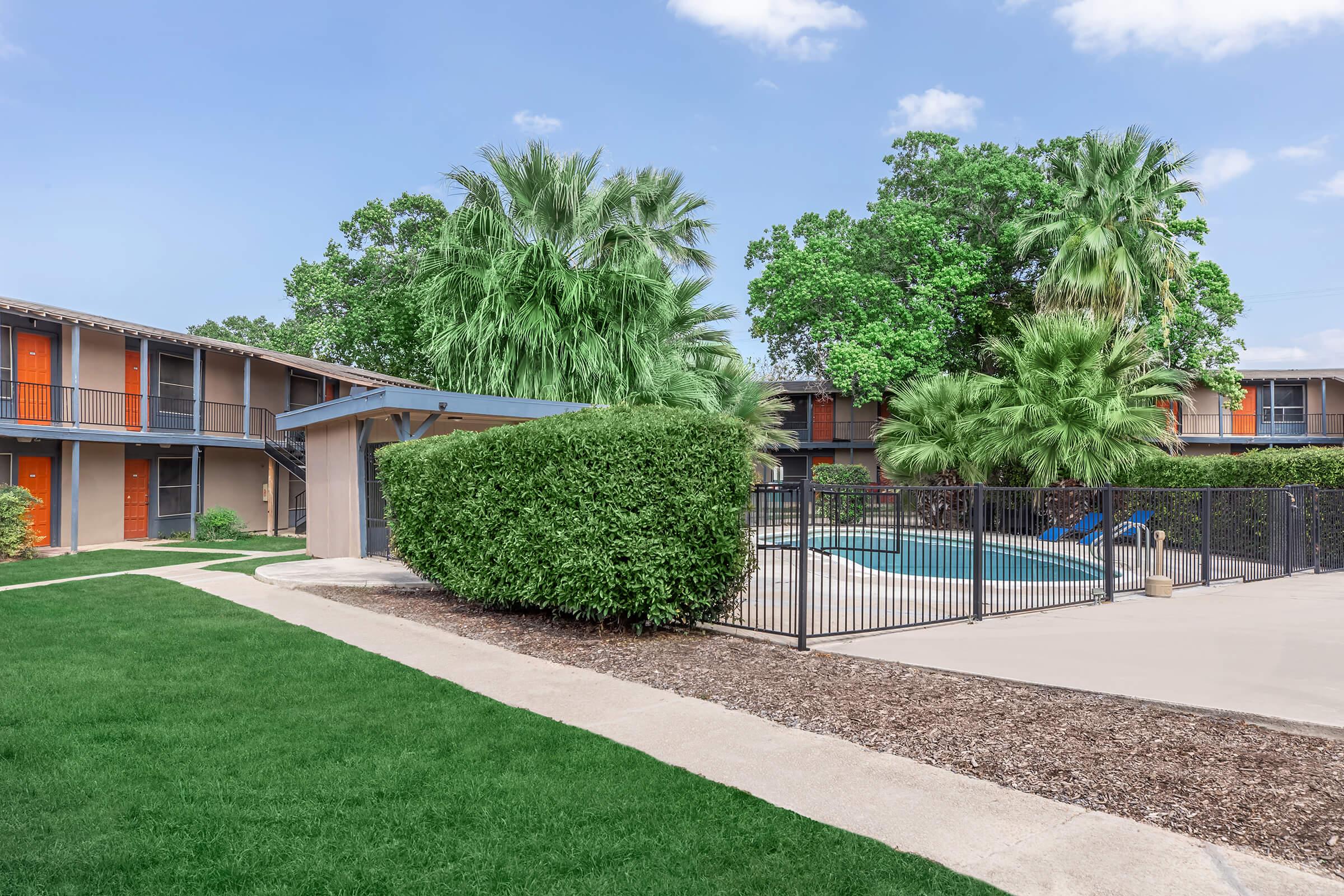 A courtyard view of a multi-story apartment complex featuring green grass, palm trees, and a fenced-in pool area surrounded by shrubs. The building has orange doors and a walkway leading to the pool. Bright blue skies are visible above, suggesting a sunny day.
