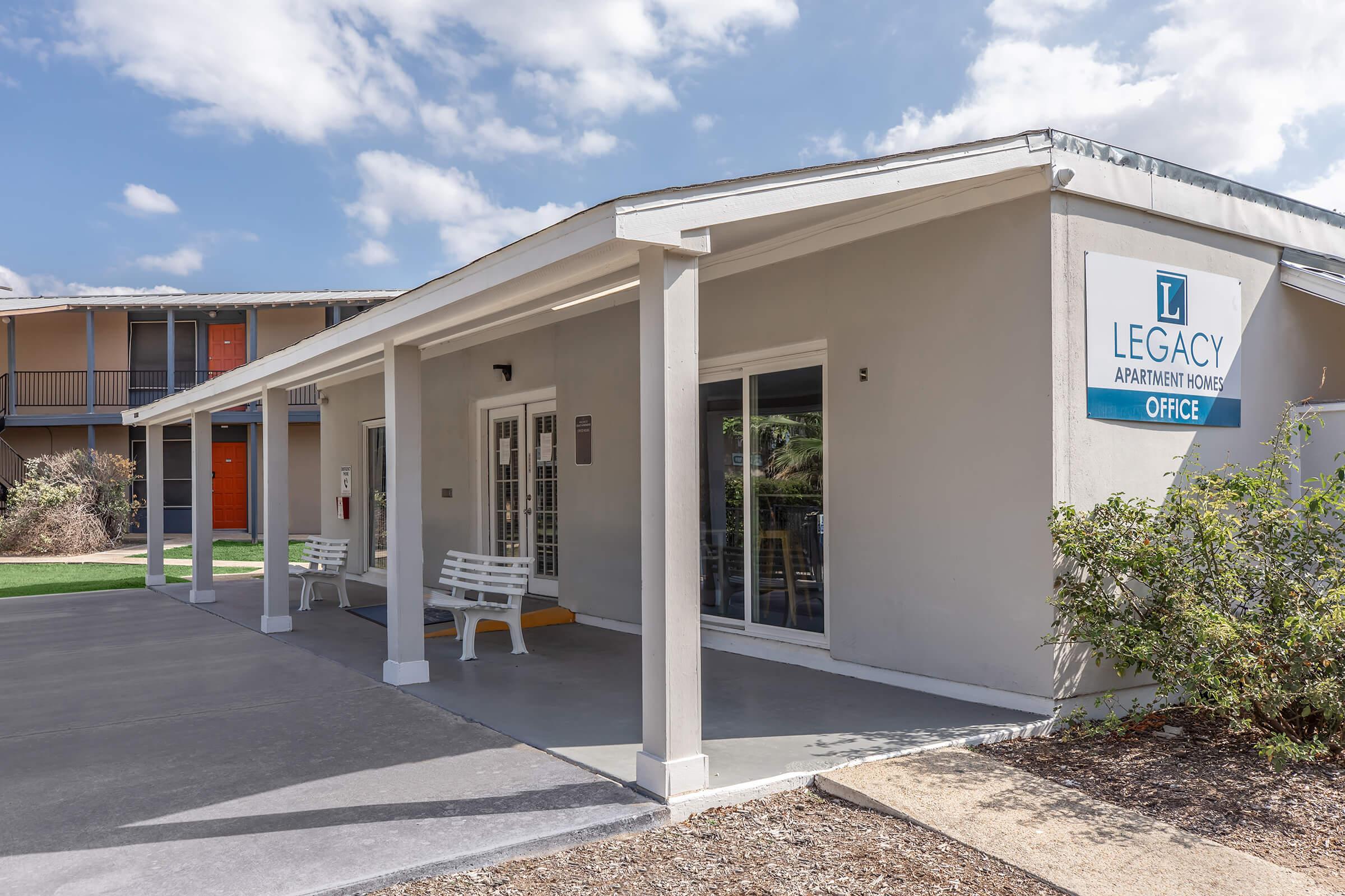Exterior view of the Legacy Apartment Homes office, featuring a modern building with a covered porch, white benches, and large windows. The office is painted in light colors, and the sign prominently displays the name. In the background, there are additional apartment units with orange doors and a well-maintained landscape.