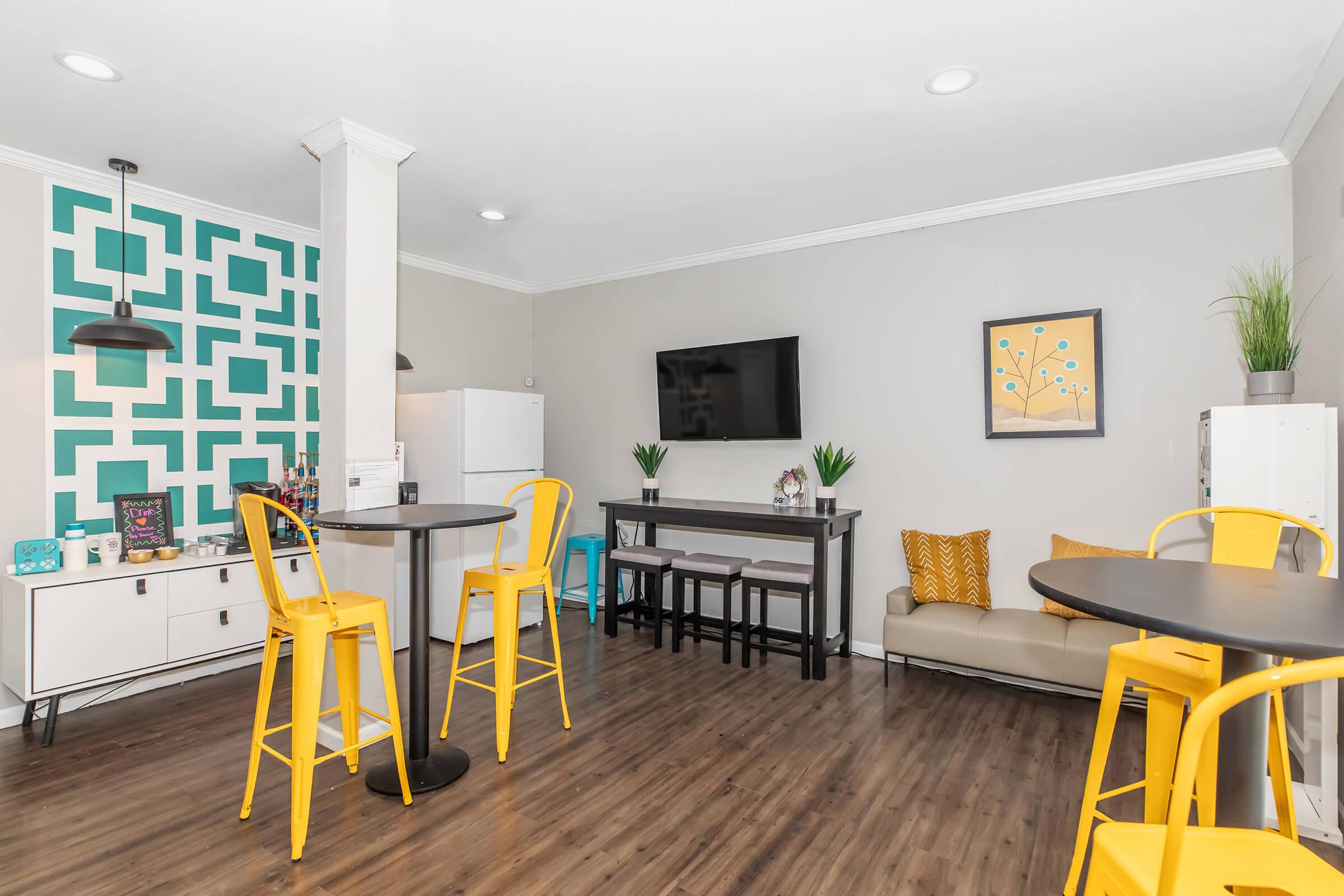 Bright and modern kitchen area featuring yellow bar stools, a refrigerator, and a wall-mounted TV. A teal geometric patterned accent wall adds style, complemented by a cozy couch and small dining tables. Decorative plants and artwork enhance the inviting atmosphere.