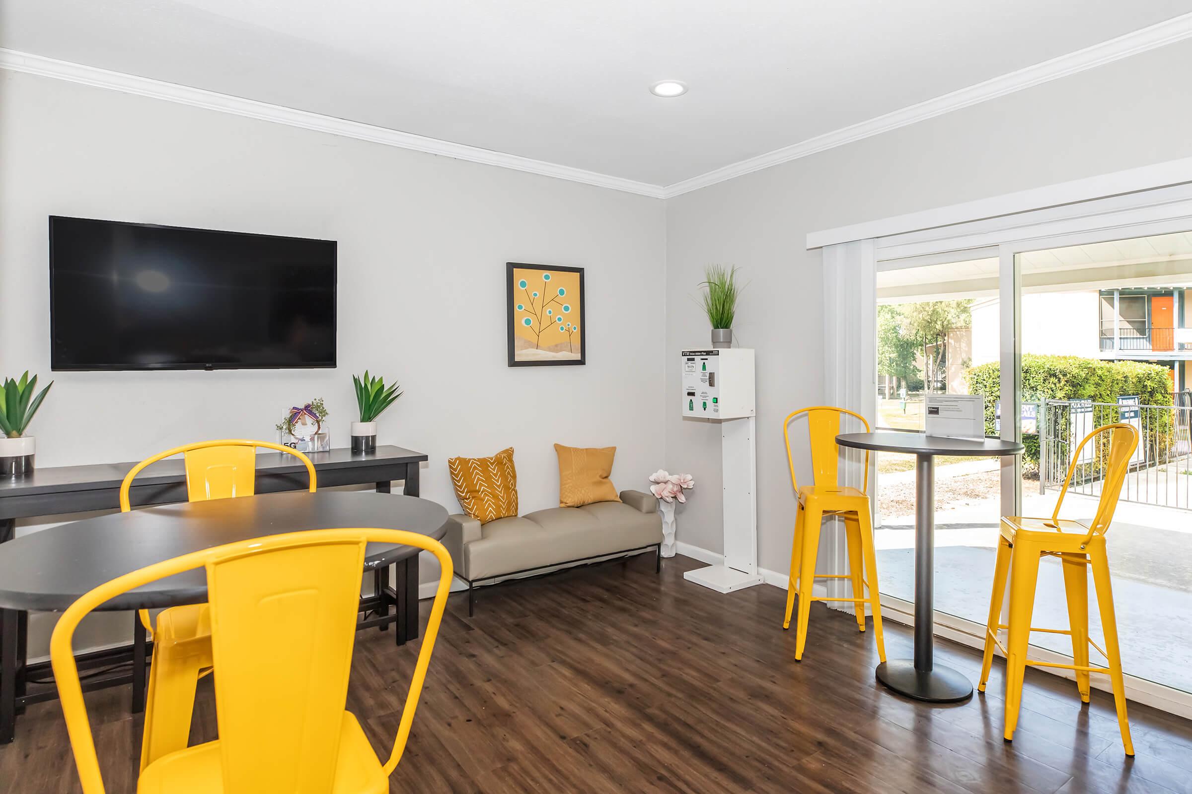 A modern lounge area featuring a gray sofa with yellow cushions, a black table with yellow chairs, and a wall-mounted TV. Green plants decorate the space, and a large window offers a view outside. Art on the wall adds a pop of color. The atmosphere is bright and inviting.