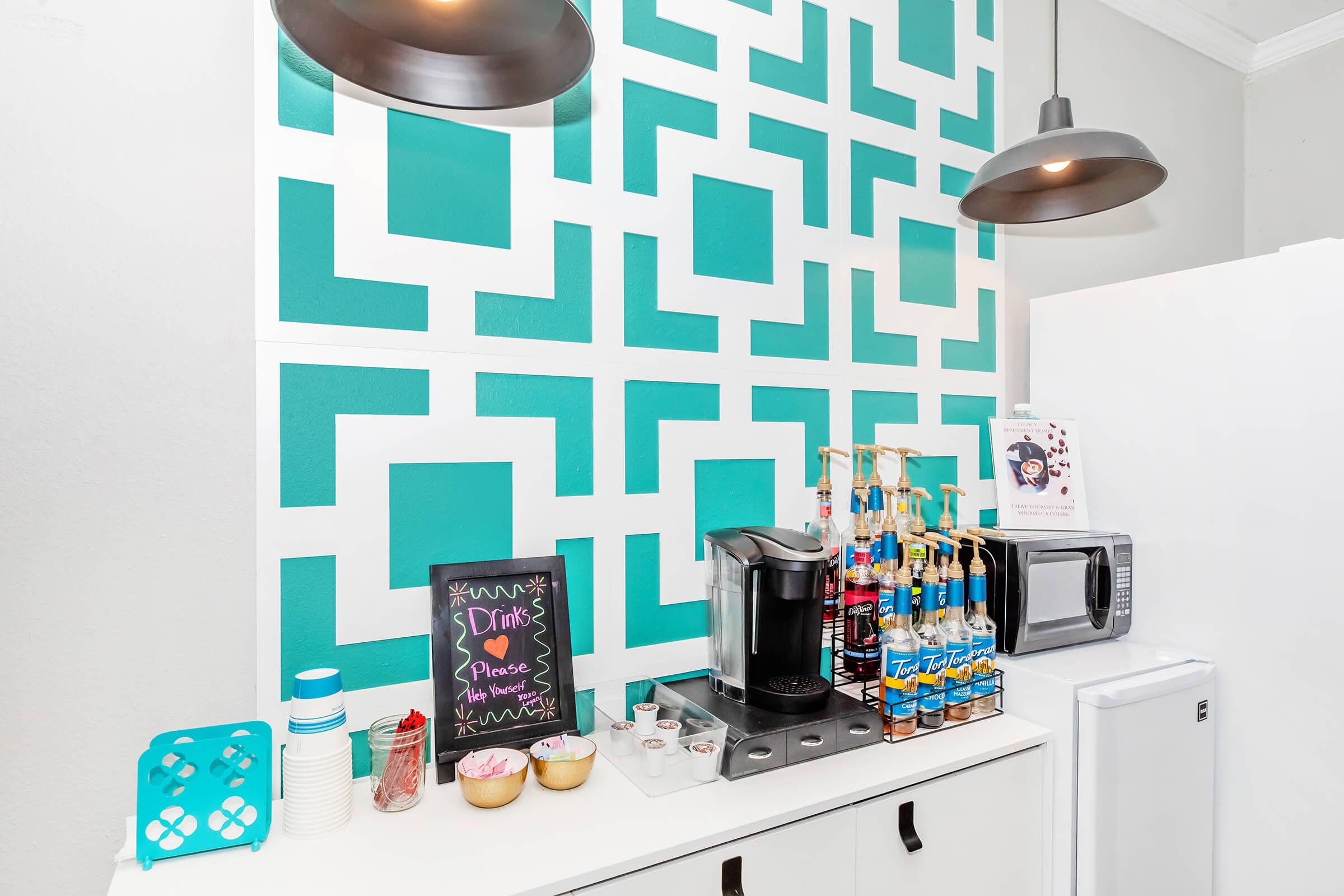 A modern kitchen corner featuring a distinctive turquoise and white patterned wall. There's a coffee maker, various colorful drink syrups, and a chalkboard sign. A microwave is visible alongside a white refrigerator. The scene is well-lit with stylish pendant lights.