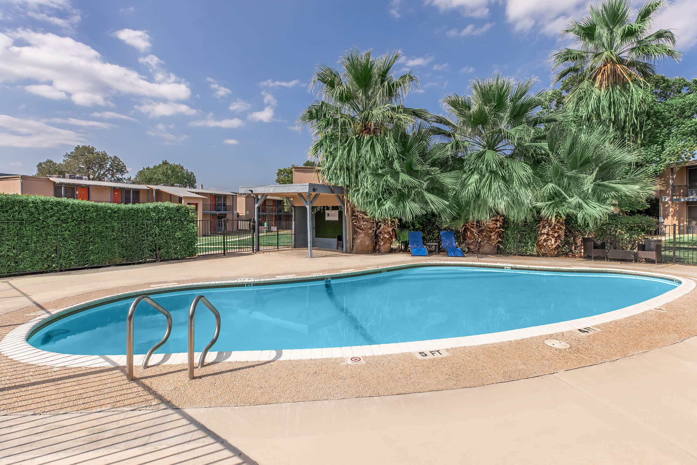 A clear swimming pool surrounded by palm trees and lush greenery. A fence encloses the area, and two blue lounge chairs are placed beside the pool. The sky is partly cloudy, and apartment buildings are visible in the background.