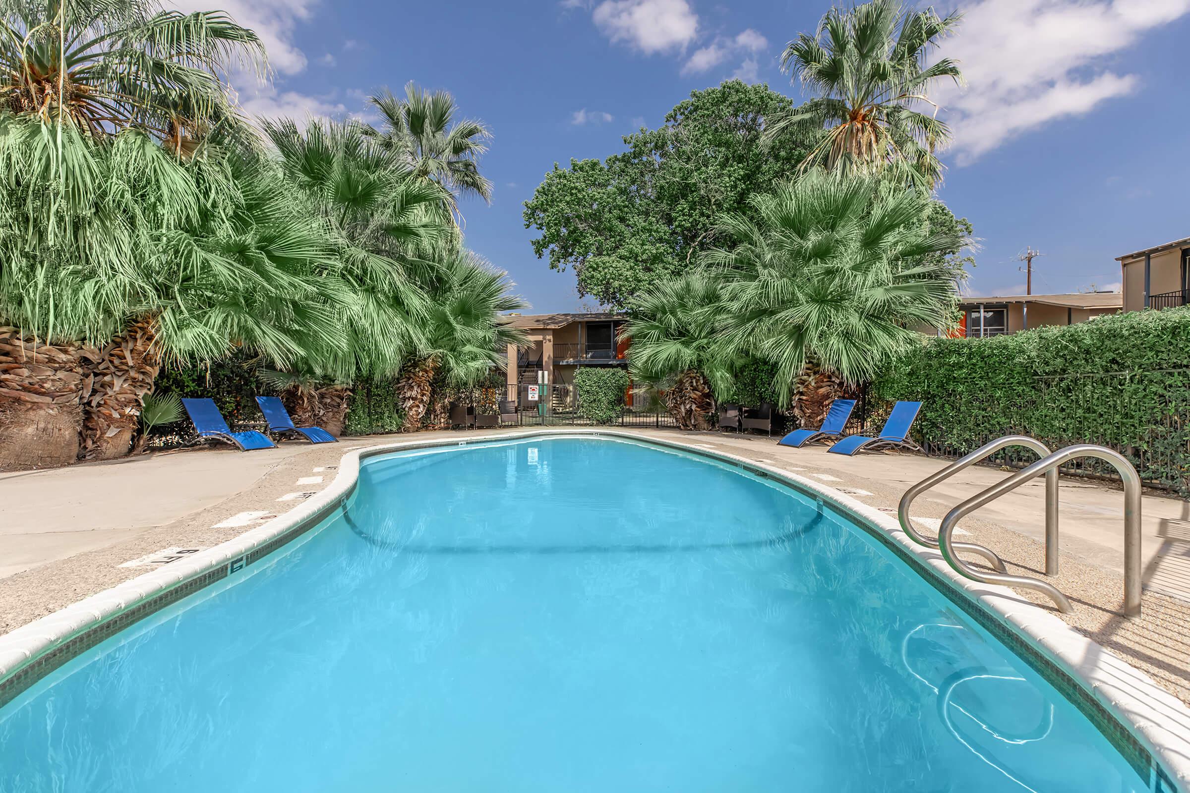 A clear blue swimming pool surrounded by lush palm trees and greenery. Sun loungers are placed on the pool deck, creating a relaxing atmosphere. The sky is bright with a few clouds, and nearby apartment buildings are visible in the background.