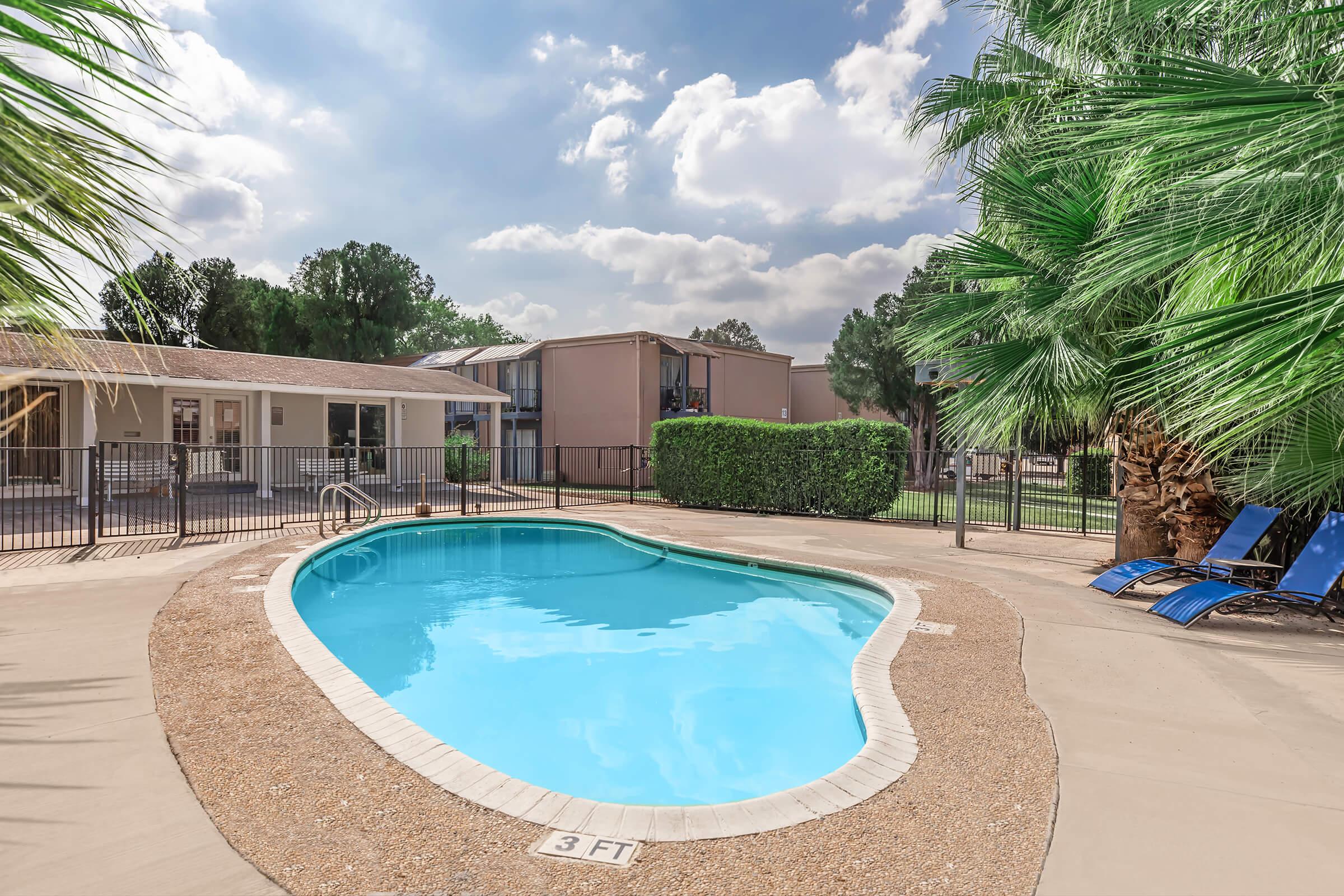 A clear blue swimming pool surrounded by a sandy area, with tropical palm trees nearby. In the background, there are two buildings with balconies and green lawns. Loungers are positioned beside the pool, under a partly cloudy sky. The setting is tranquil and inviting, perfect for relaxation.