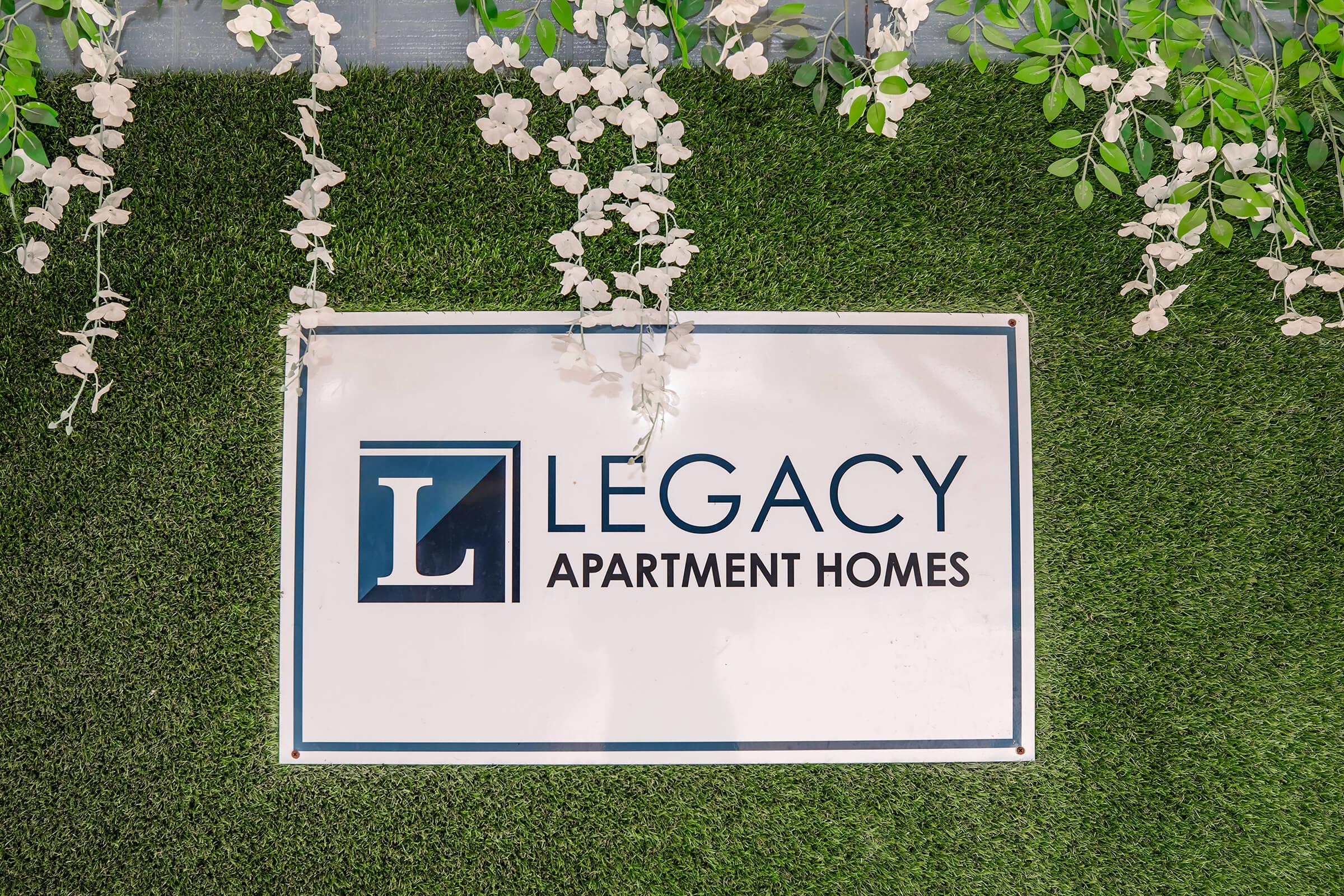 Sign for "Legacy Apartment Homes" displayed against a green artificial grass background, decorated with white floral elements. The logo features a prominent "L" in blue.