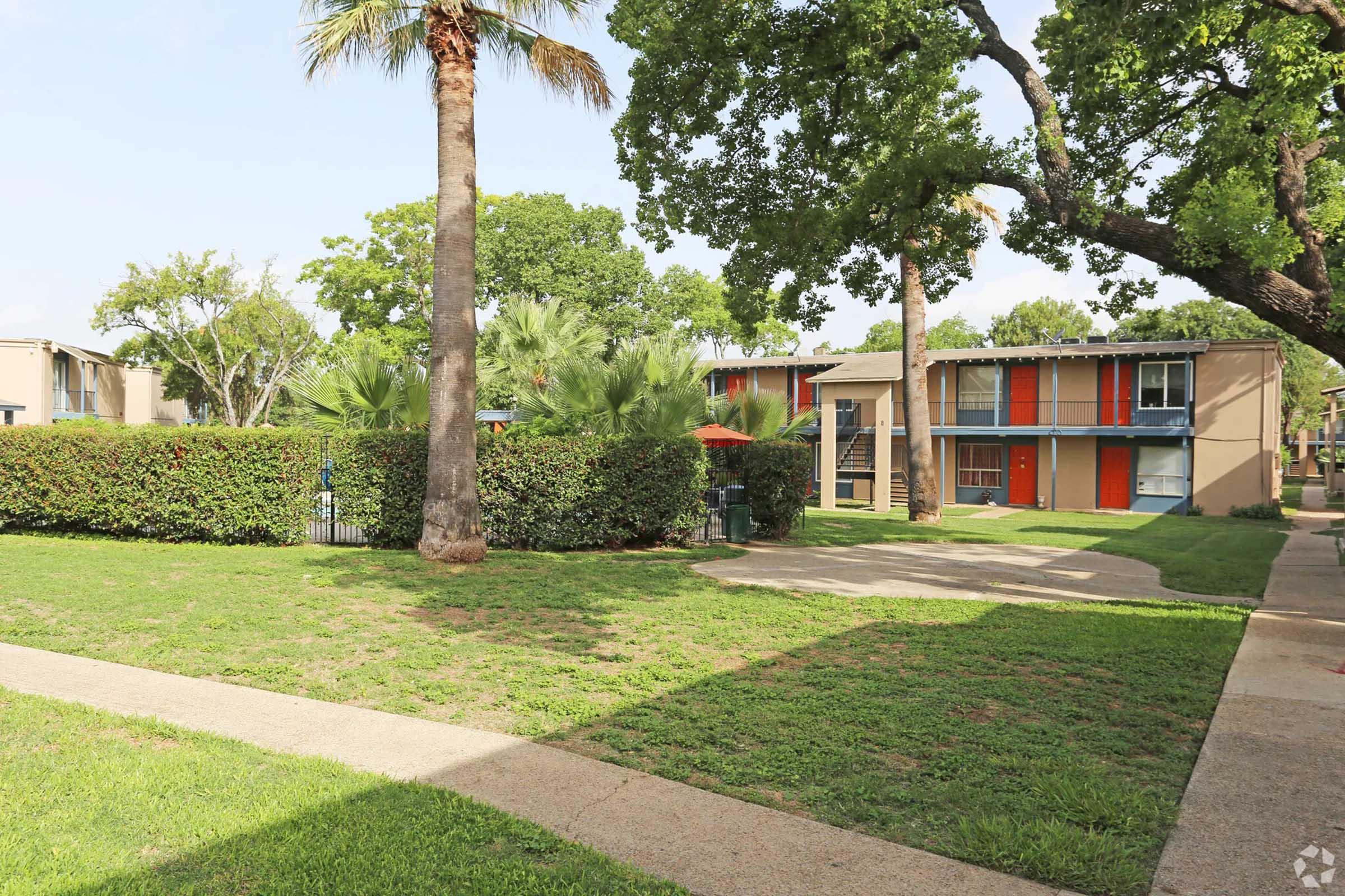 A grassy courtyard with neatly trimmed hedges and palm trees, featuring two buildings with colorful exteriors, one orange and one blue. A pathway cuts through the area, leading to the buildings. An umbrella is visible near the hedges, adding a relaxed atmosphere to the scene.