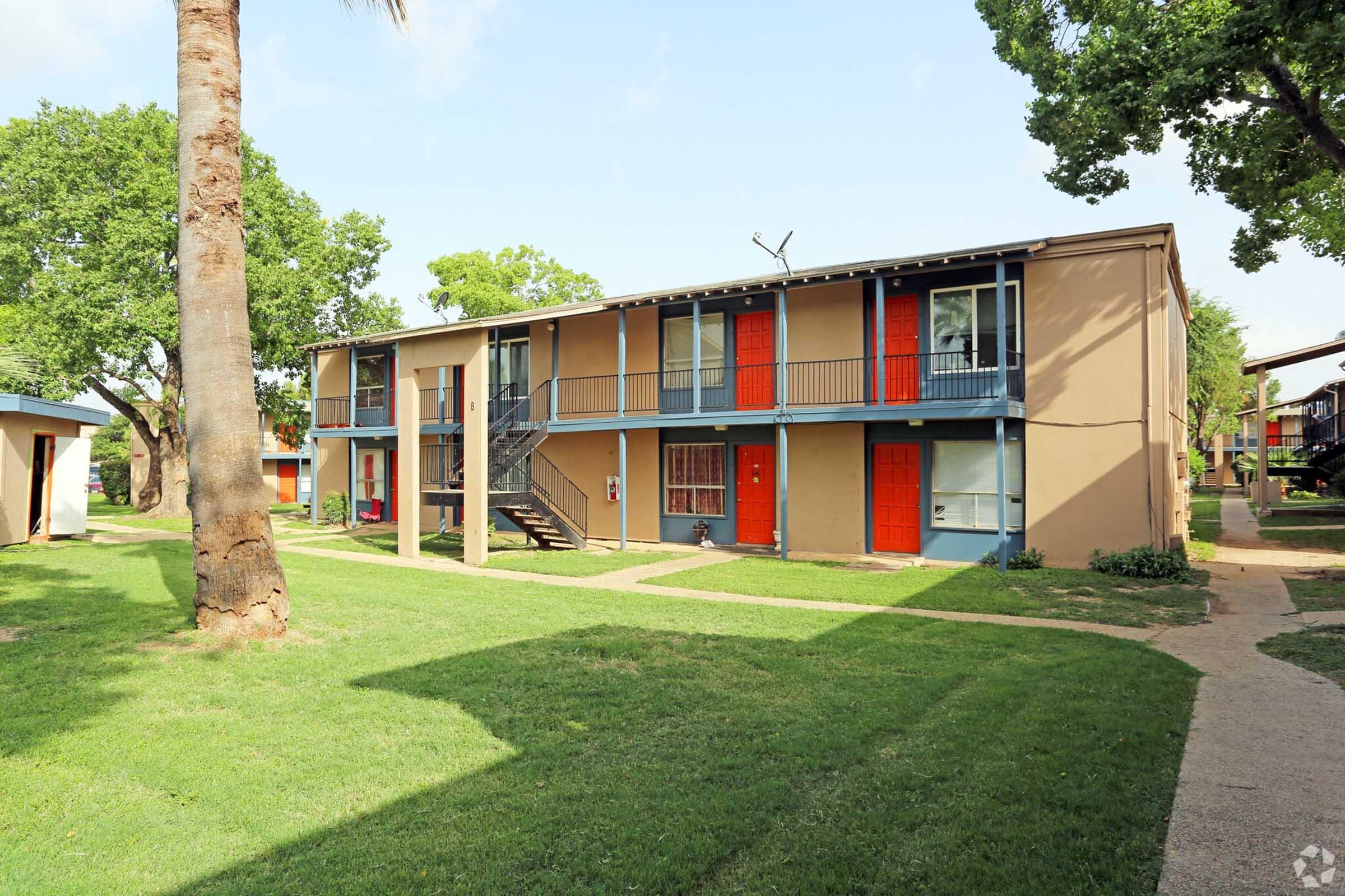 Two-story apartment building set in a green landscape with trees. The exterior features beige walls with bright orange doors. A pathway winds through the grass, leading to the entrances of the units. The scene is sunny with a clear blue sky.