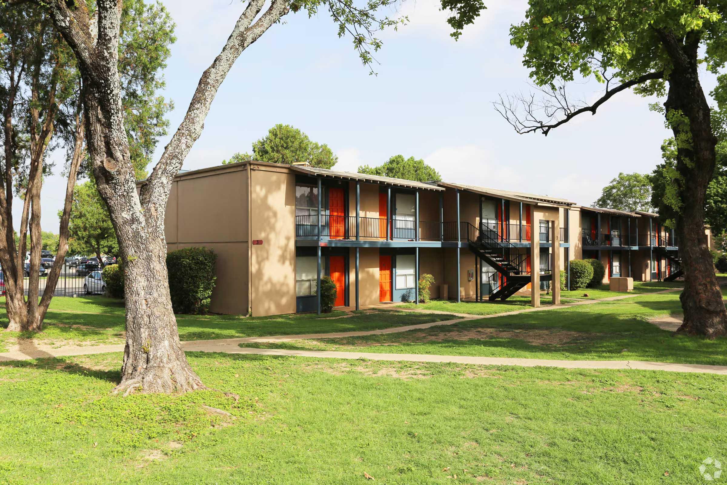 A two-story apartment complex featuring a beige exterior with orange doors and blue railings. The building is surrounded by greenery, including trees and grass, with a pathway leading to the entrance. The sky is partly cloudy, and parking is visible in the background.