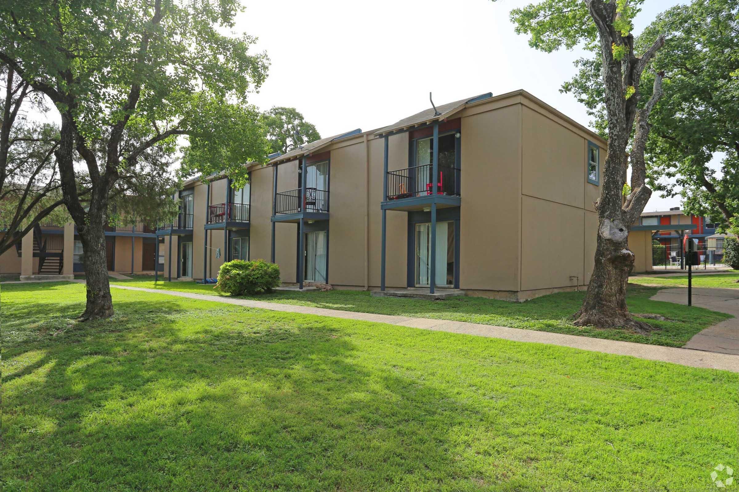 A view of two-story apartment buildings surrounded by grassy lawns and trees. The buildings have balconies, and the area appears well-maintained with clear skies.