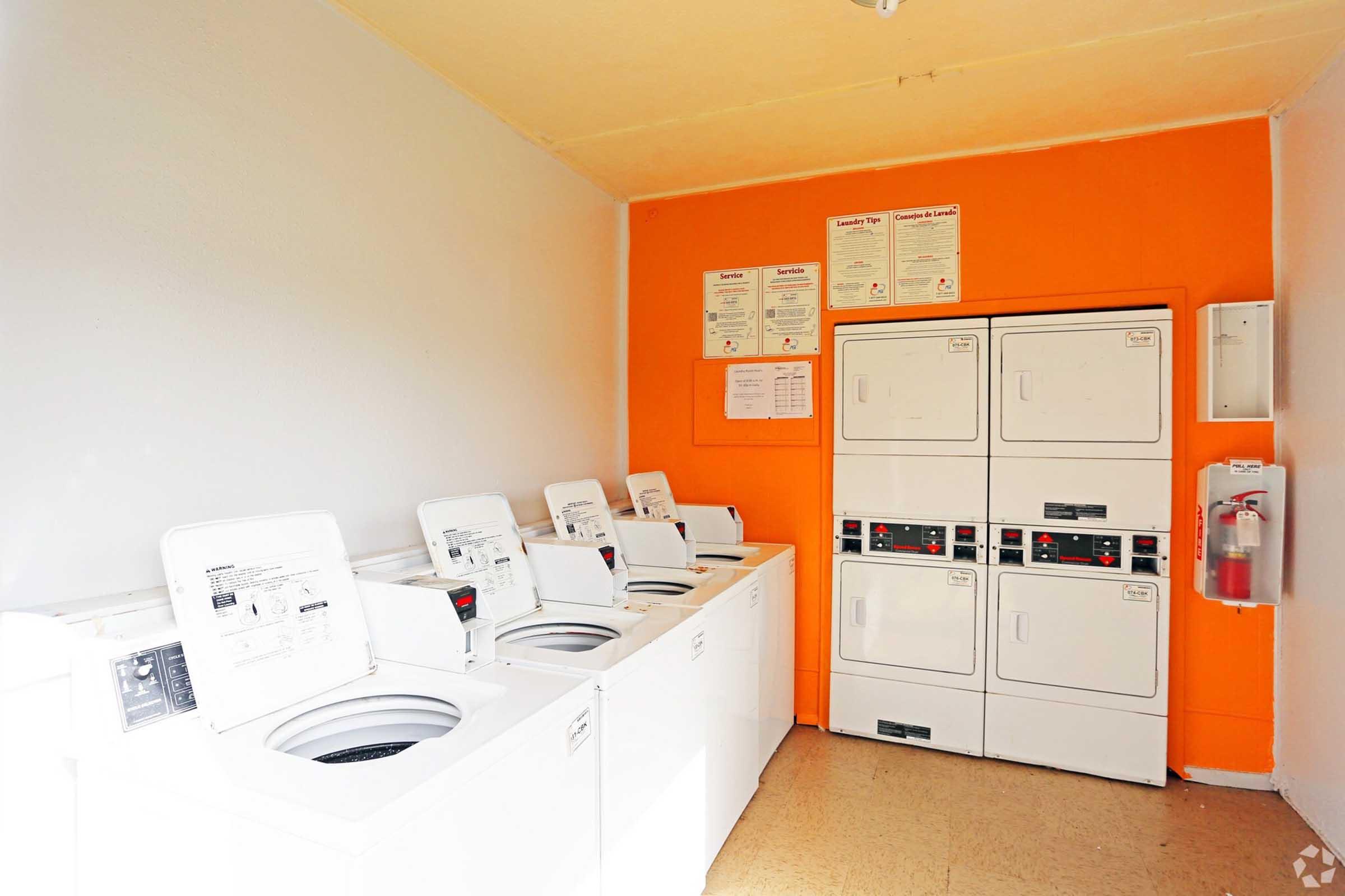 A laundry room featuring several white washing machines and stacked dryers against an orange wall. Informational posters are displayed on the wall above the machines, and a fire extinguisher is mounted nearby. The floor is made of light-colored tiles.