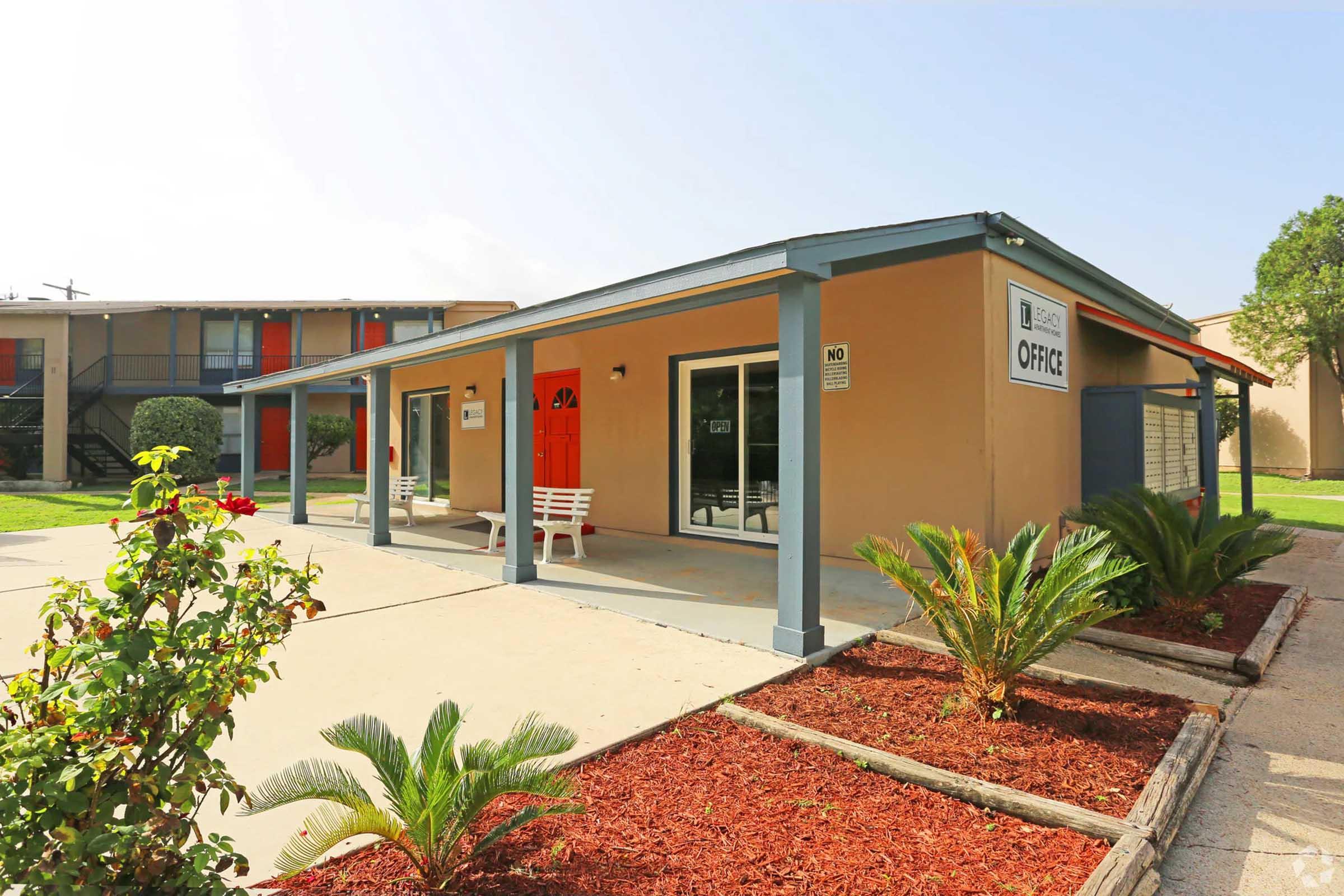 A single-story office building with a covered porch, featuring large windows and a sign indicating "OFFICE." In front, there are two white benches and decorative landscaping with palm plants and red mulch. The surrounding area includes another building in the background and a well-maintained lawn.