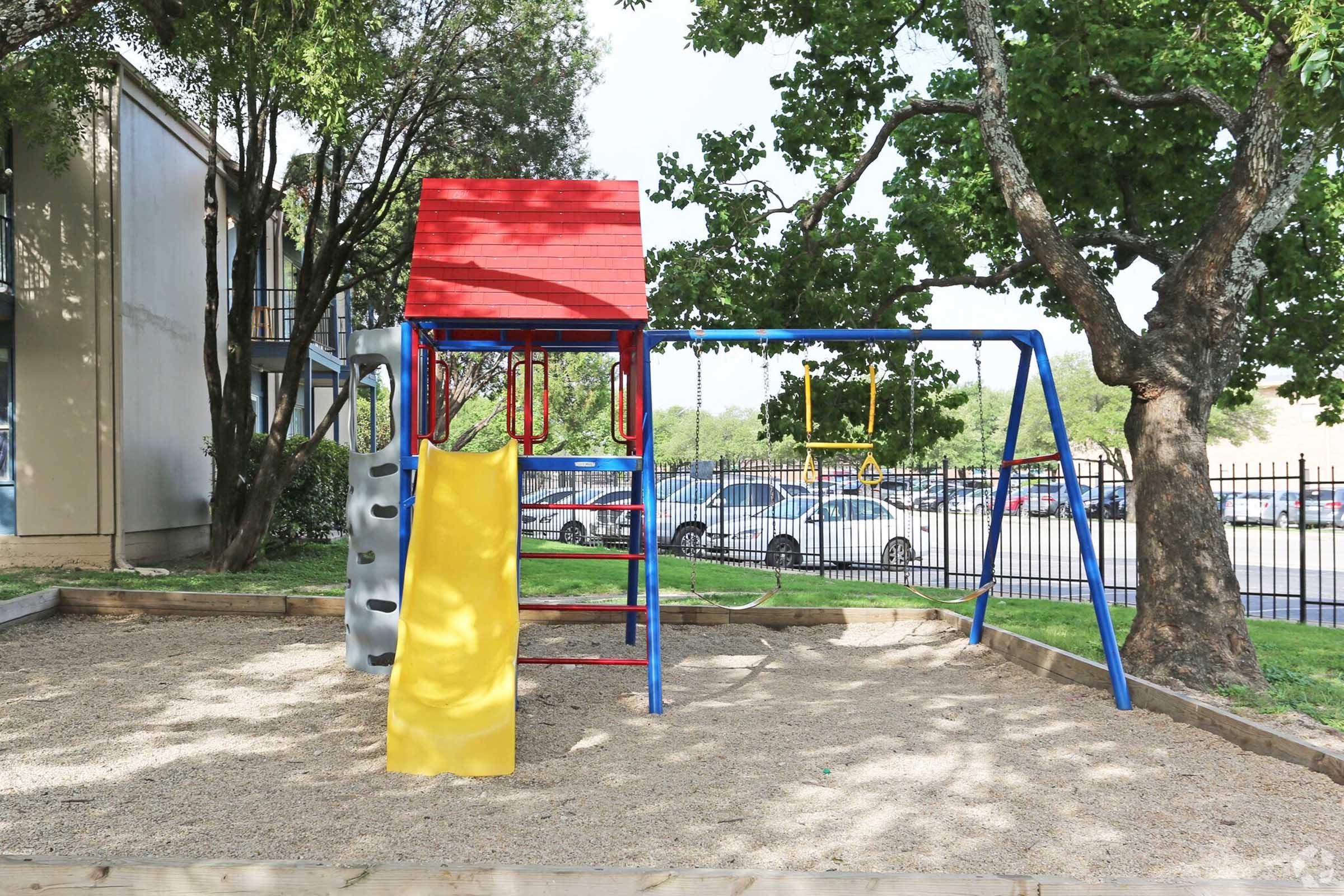 A playground featuring a colorful climbing structure with a red roof, slide, and swings, set in a sandy area surrounded by trees. In the background, there are parked cars and a fence.