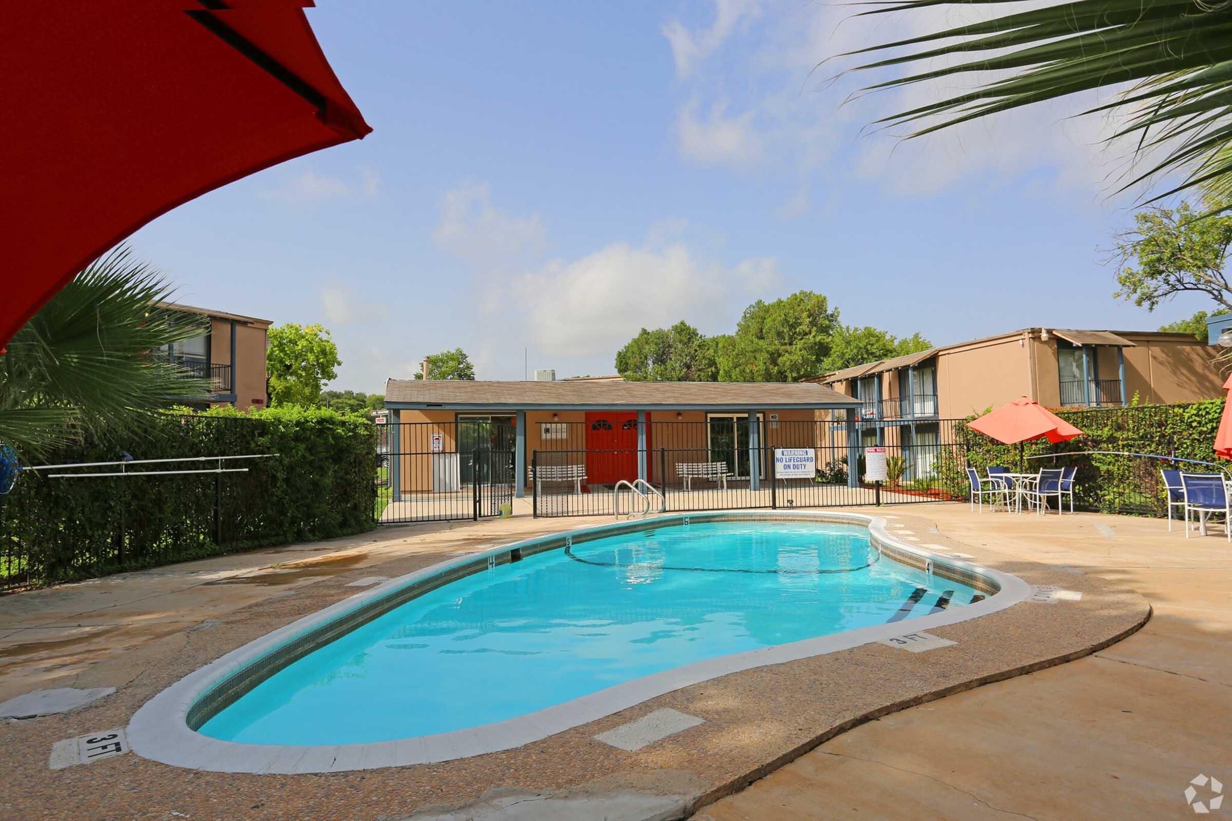 A swimming pool surrounded by lounge chairs and greenery, with a fence and buildings in the background. Bright blue sky and a few clouds are visible, with red umbrellas providing shade near the pool area.