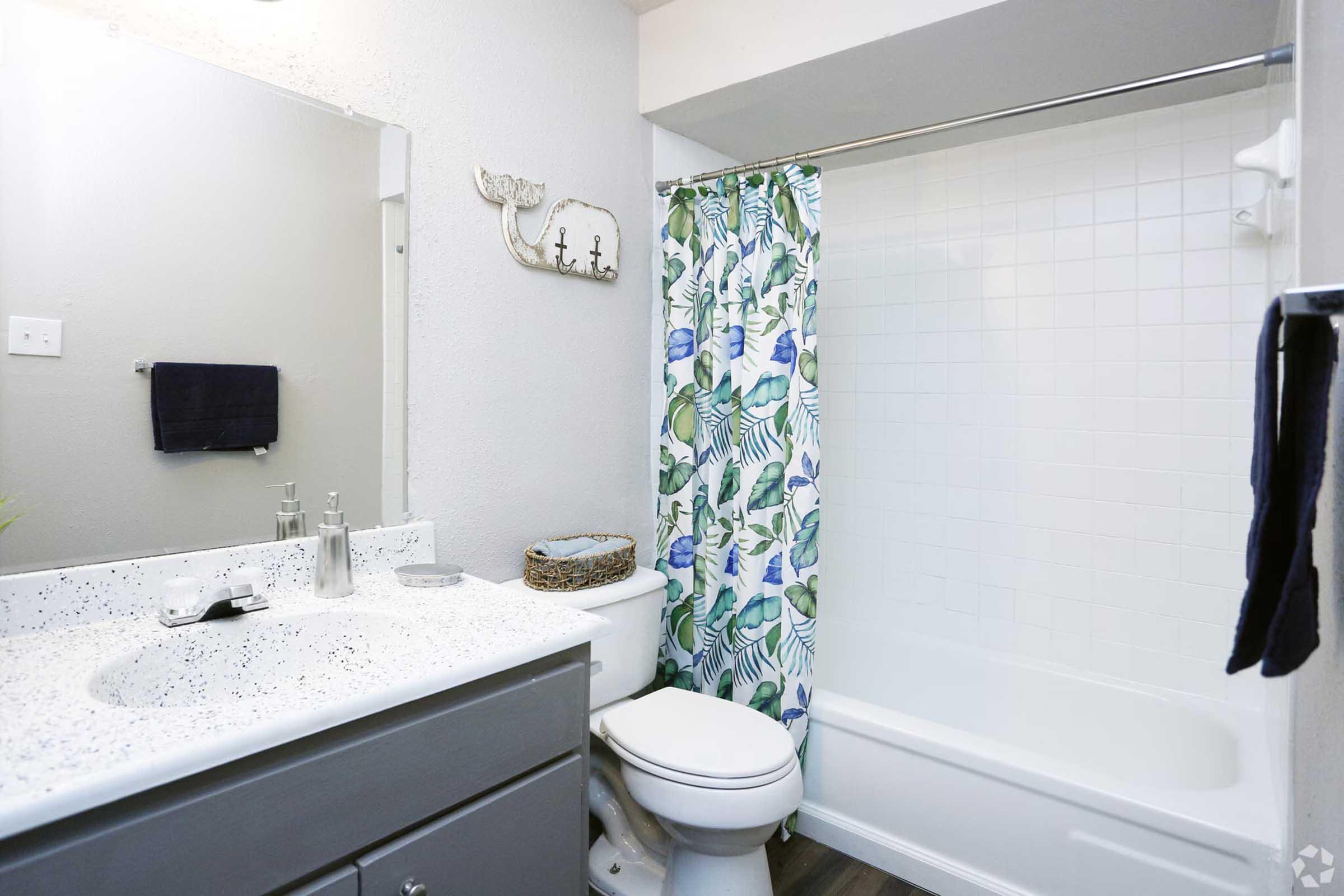 A clean and modern bathroom featuring a white bathtub with a shower curtain decorated with green leaves and blue flowers. The vanity has a speckled countertop with silver fixtures and a mirror above. A towel hangs from a rack nearby, and there are plants and toiletries neatly arranged.