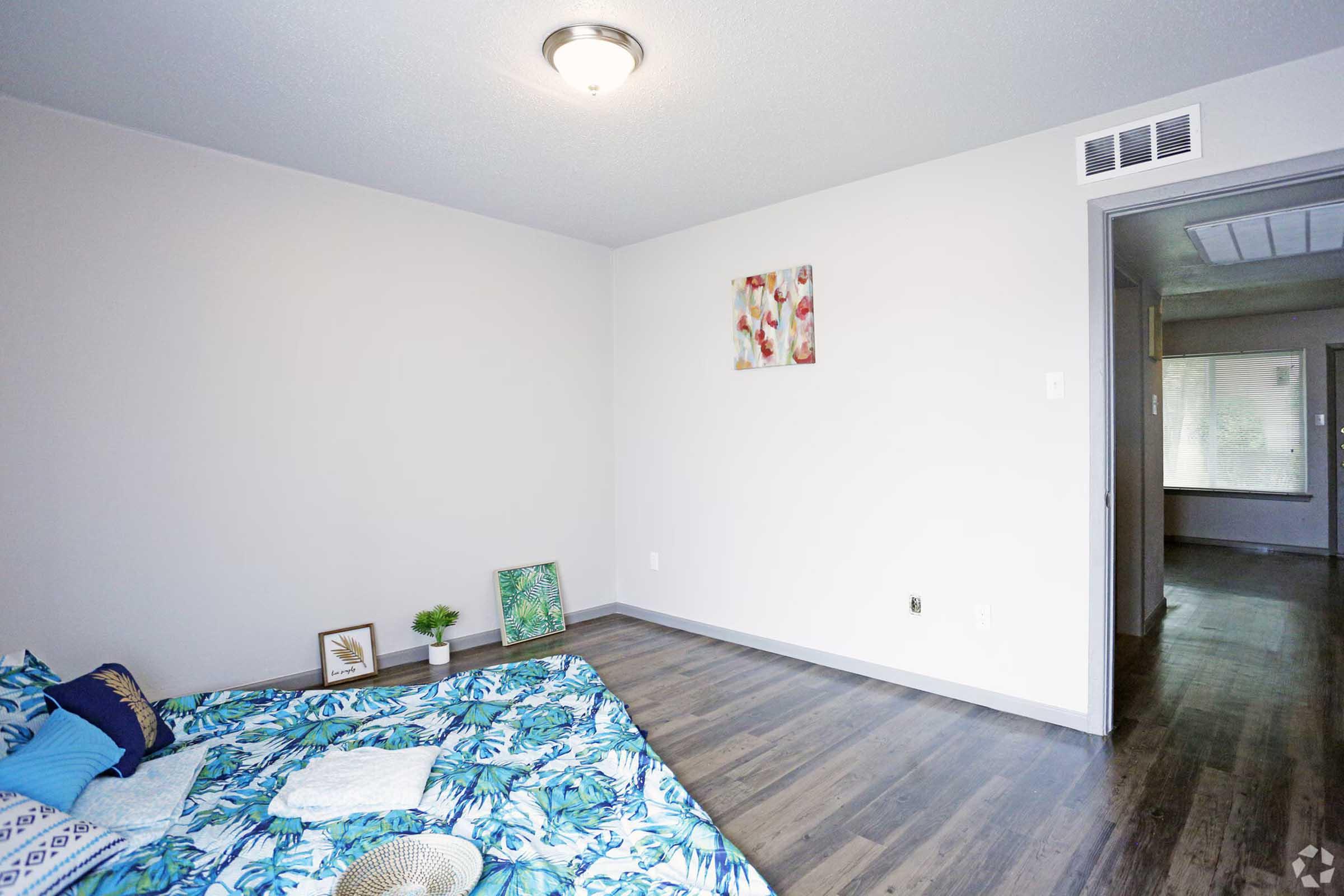 A simple and modern bedroom featuring a floral-themed bedspread, a small decorative plant, and two framed pieces of artwork on the wall. The room has light-colored walls, wood-like flooring, and a doorway leading to another room, creating a spacious and inviting atmosphere.