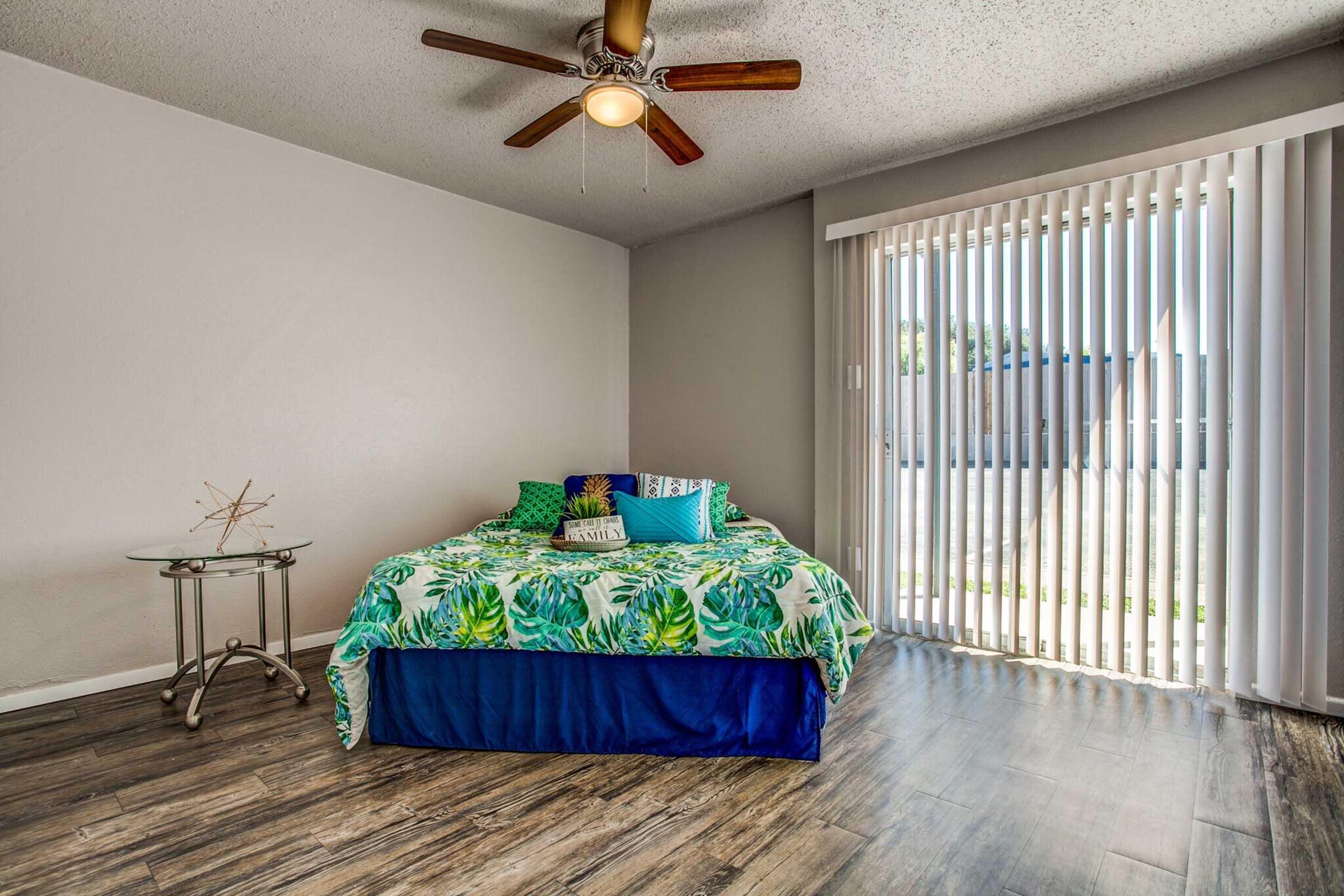 A cozy bedroom featuring a bed with a colorful tropical-themed blanket and decorative cushions. The room has wood-look flooring, a ceiling fan, and large sliding glass doors that lead to an outdoor area with vertical blinds, allowing natural light to enter. A small side table is next to the bed.