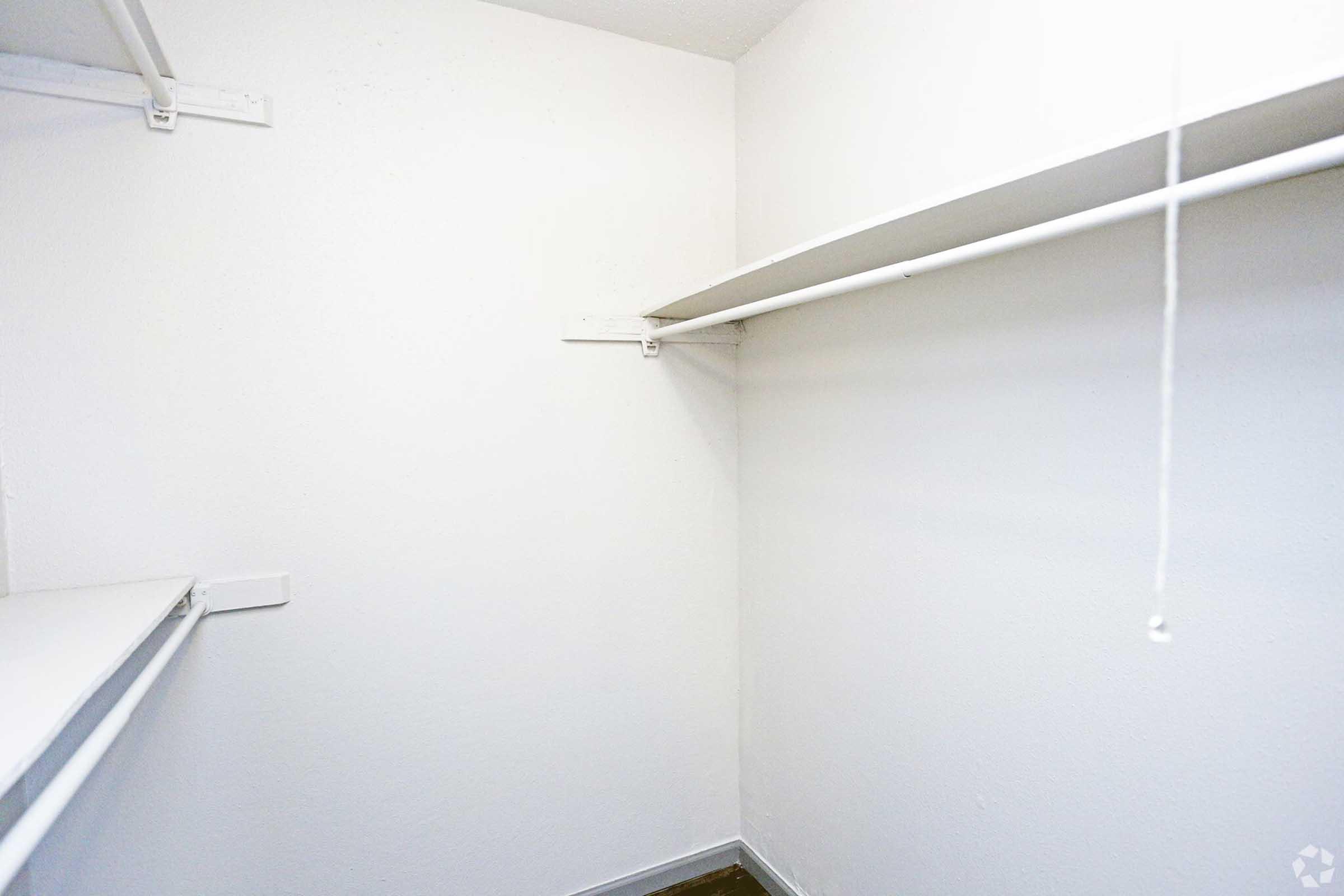 Empty closet with white walls and two shelves. The shelves are made of metal and positioned at different heights. There is a pull string hanging from the ceiling, indicating a light fixture. The floor appears dark, contrasting with the bright walls and shelves.