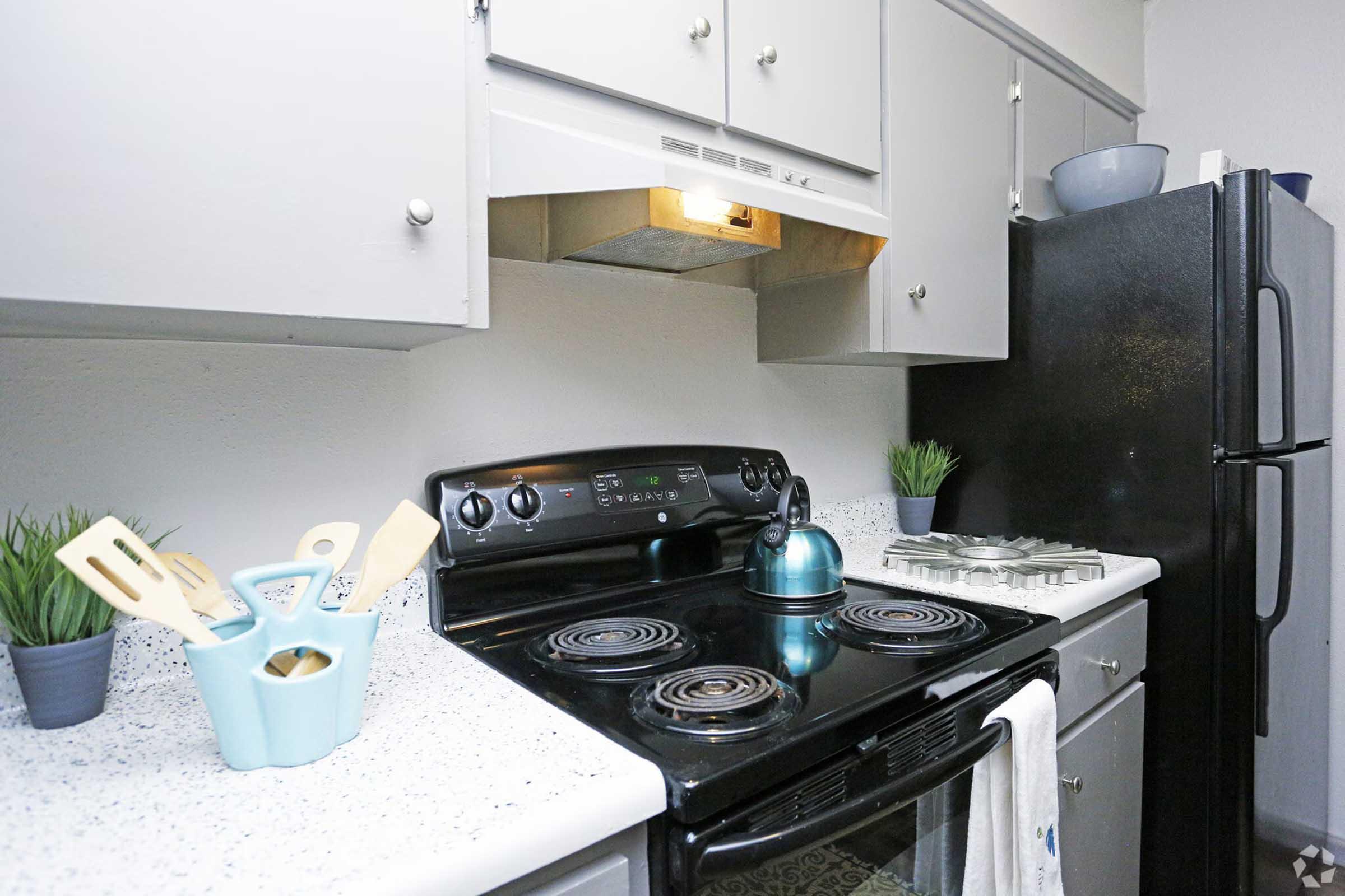 A kitchen scene featuring a black stove and oven with a stovetop kettle, a microwave above, and a black refrigerator. Light gray cabinets are above the stove, accompanied by a small potted plant and kitchen utensils in a blue container on the counter. The countertop has a speckled pattern, adding a modern touch.