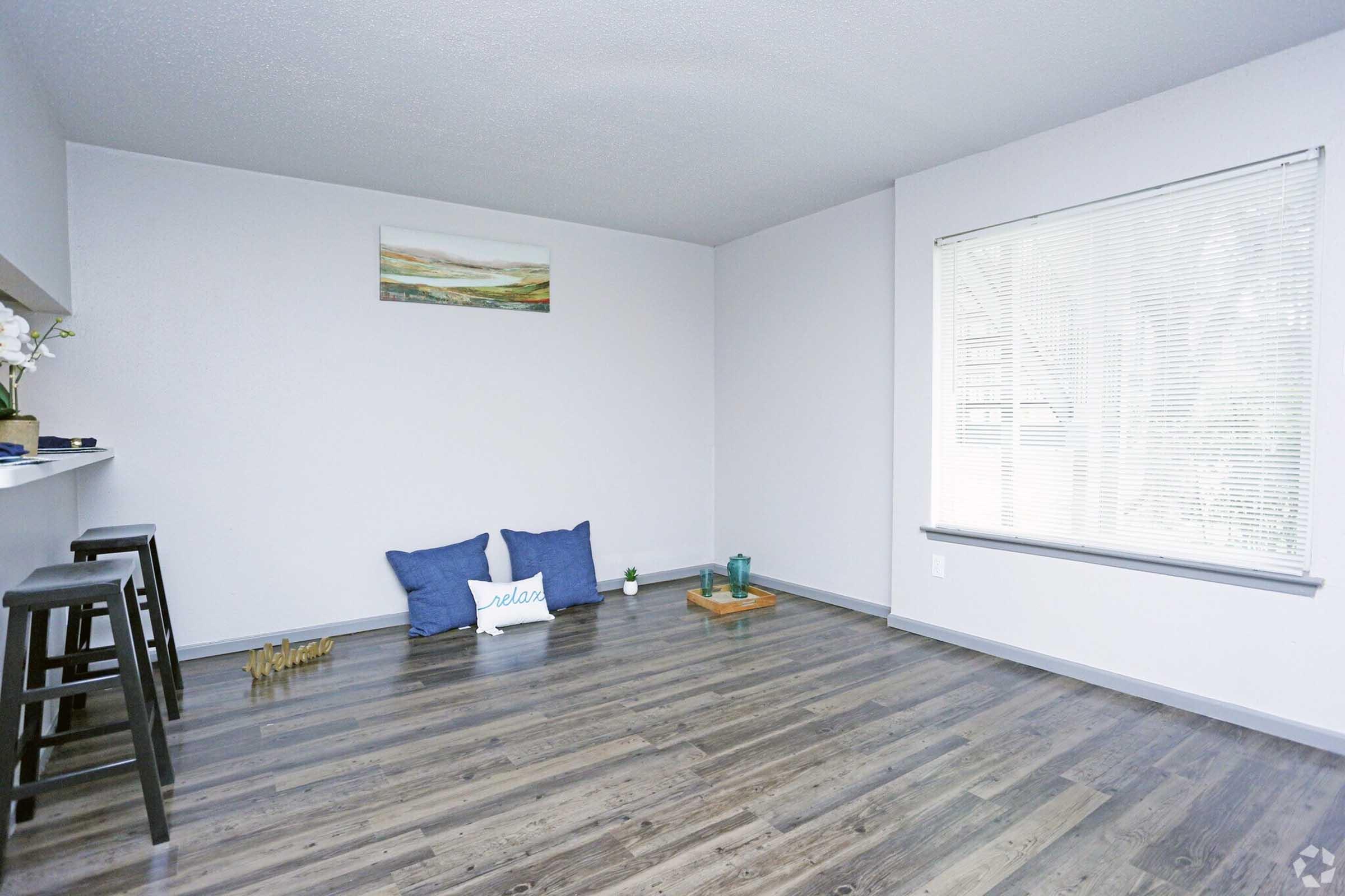 A bright, modern room with light-colored walls and dark wooden flooring. There is a small table with two chairs, a larger empty wall space, and minimal furniture, creating a spacious and uncluttered atmosphere. Natural light streams in from a window, enhancing the openness of the space.