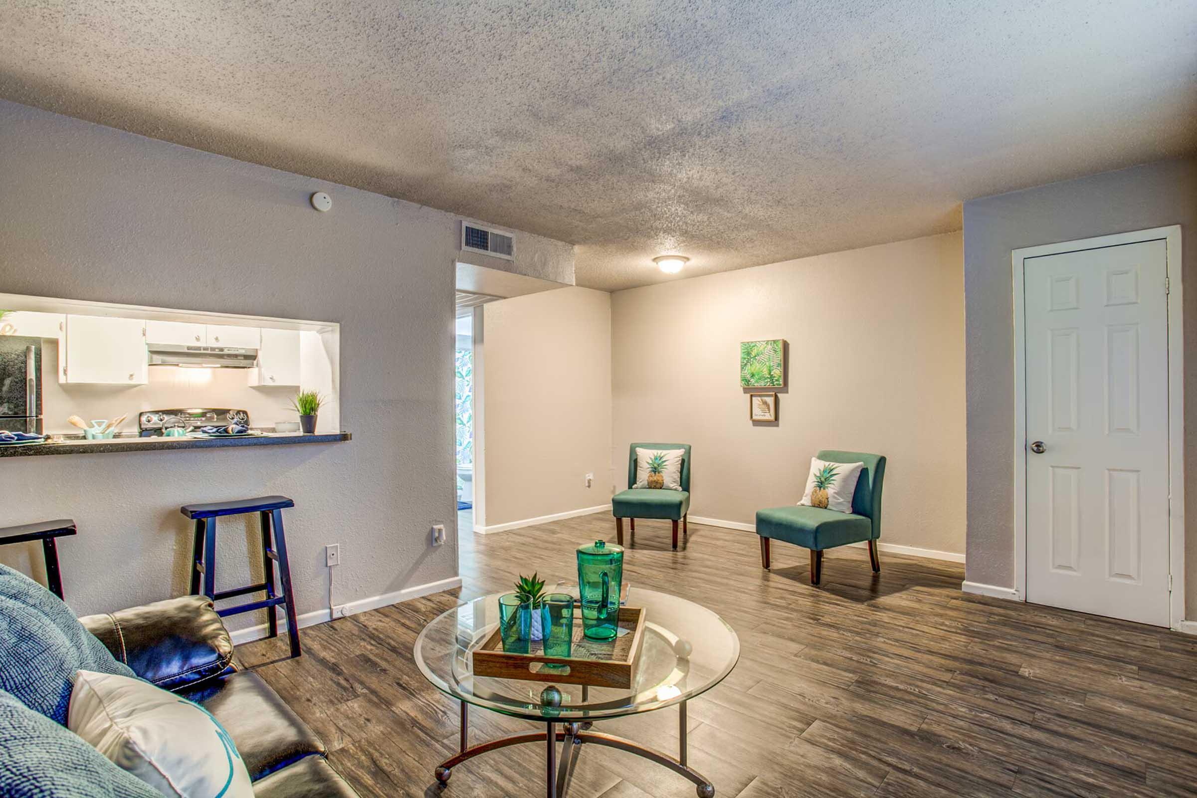 A cozy living room featuring a glass coffee table, two teal chairs, and a neutral-toned wall. The space is well-lit, with an open kitchen in the background, showcasing white cabinetry. Green decorations add a touch of color, enhancing the inviting atmosphere.