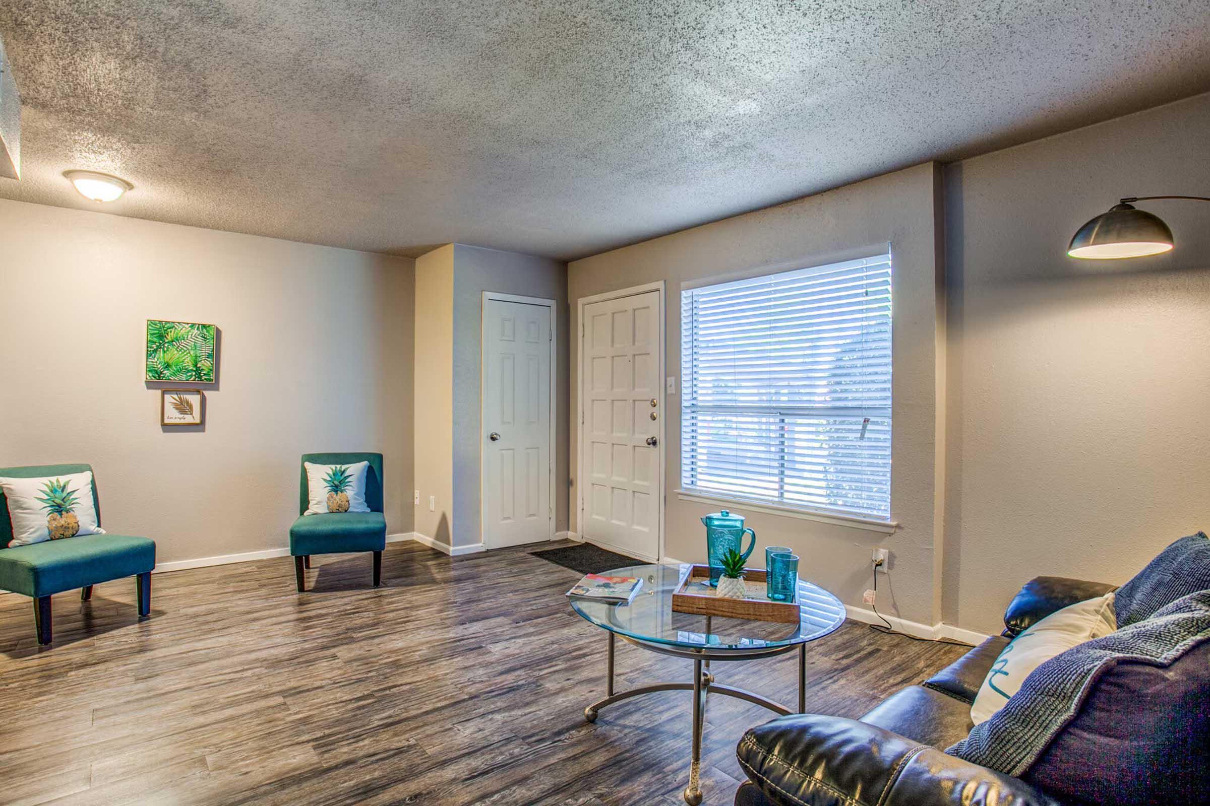 A cozy living room featuring two teal chairs with pineapple print pillows, a glass coffee table, and a comfortable black sofa. Natural light streams in through a window with white blinds. The walls are painted in neutral tones, and a small artwork of greenery is displayed on one wall.