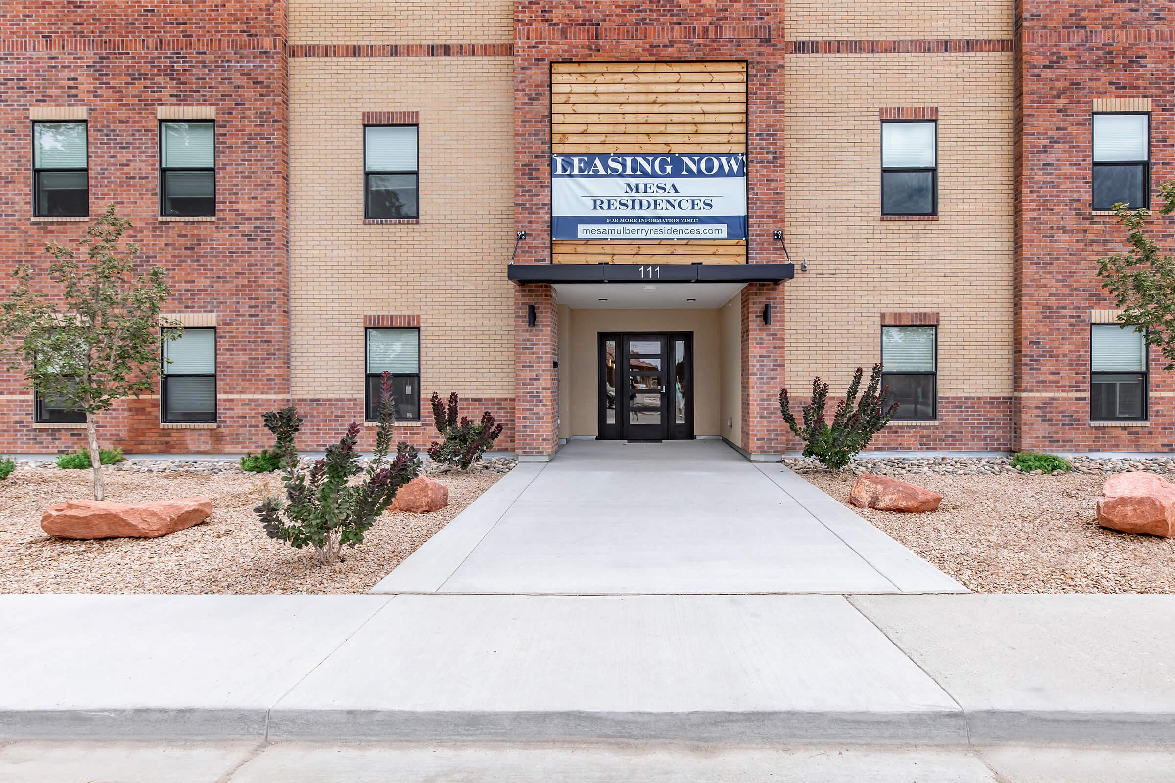 Entrance of a residential building with a sign that reads "Leasing Now" for Mesa Residences. The building features a brick facade and several windows. The path leads to the double doors, with decorative rocks and small shrubs on either side. The area is landscaped with gravel.