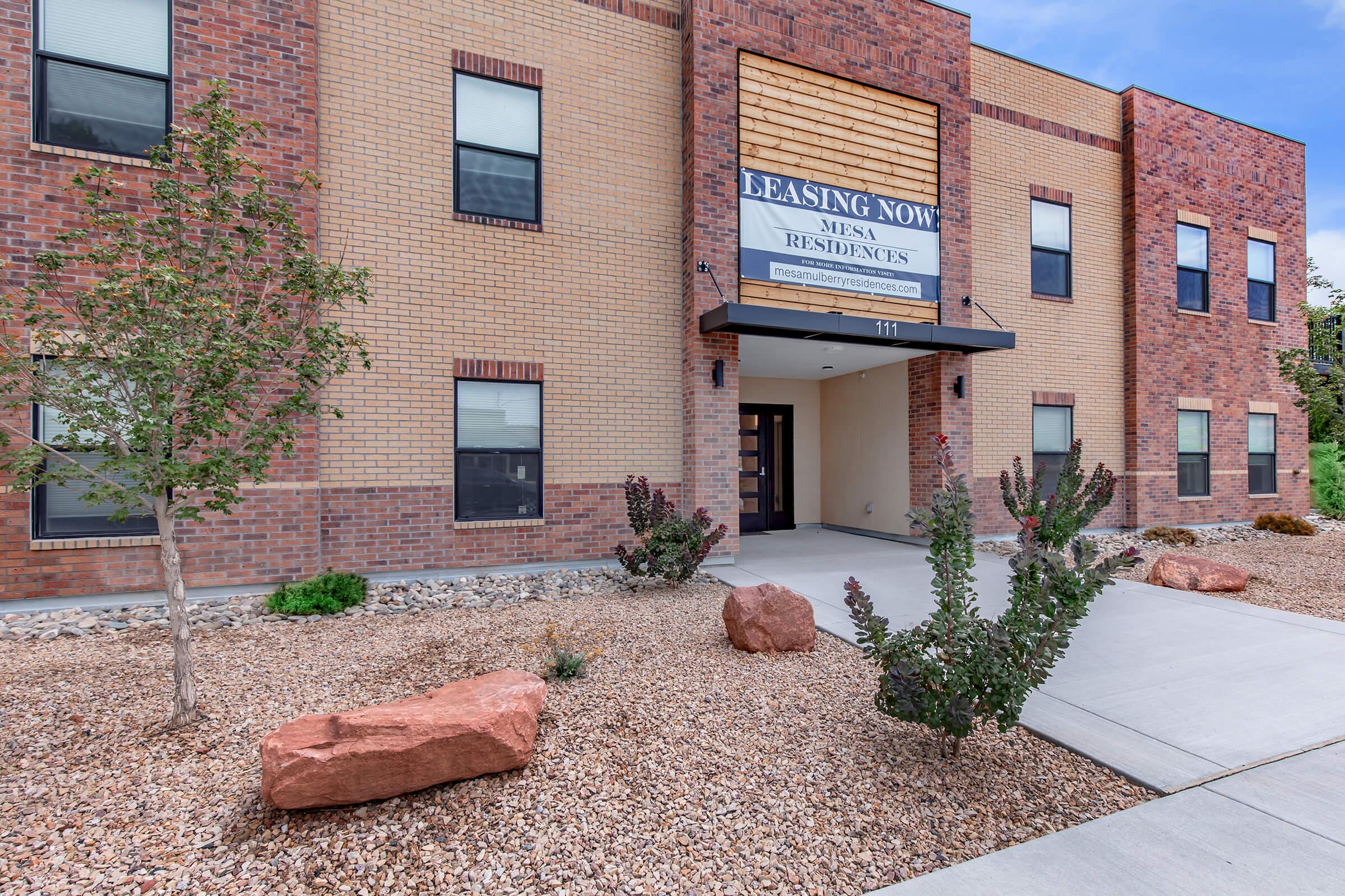 Exterior view of a modern brick building featuring a prominent sign that reads "Leasing Now" for Mesa Residences. The entrance is flanked by landscaped areas with rocks and plants, and a wide concrete pathway leads to the door. Clear blue sky in the background.