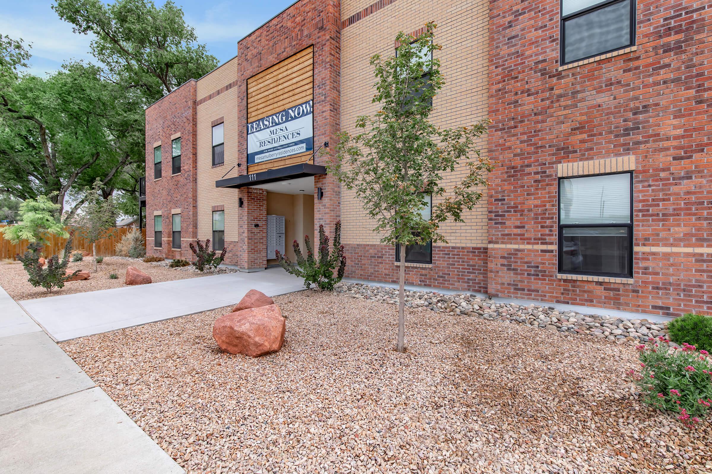 Exterior view of a residential building with a sign that reads "Leasing Now" above the entrance. The building features a mix of brick and modern architectural elements, surrounded by landscaped gravel beds and low shrubs. Large windows are visible, and there are decorative boulders in the garden area.