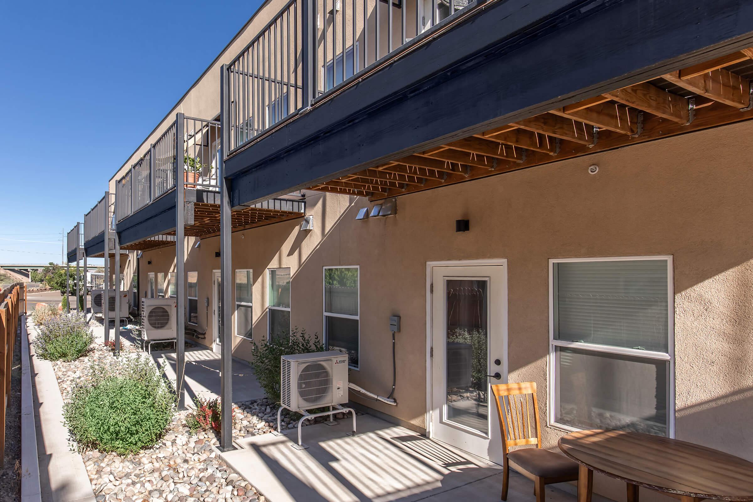 A view of an apartment building featuring multiple upper-level balconies, an outdoor seating area with a table and chair, and air conditioning units. The landscape includes decorative rocks and small shrubs, with clear blue skies overhead. The setting appears modern and well-maintained.