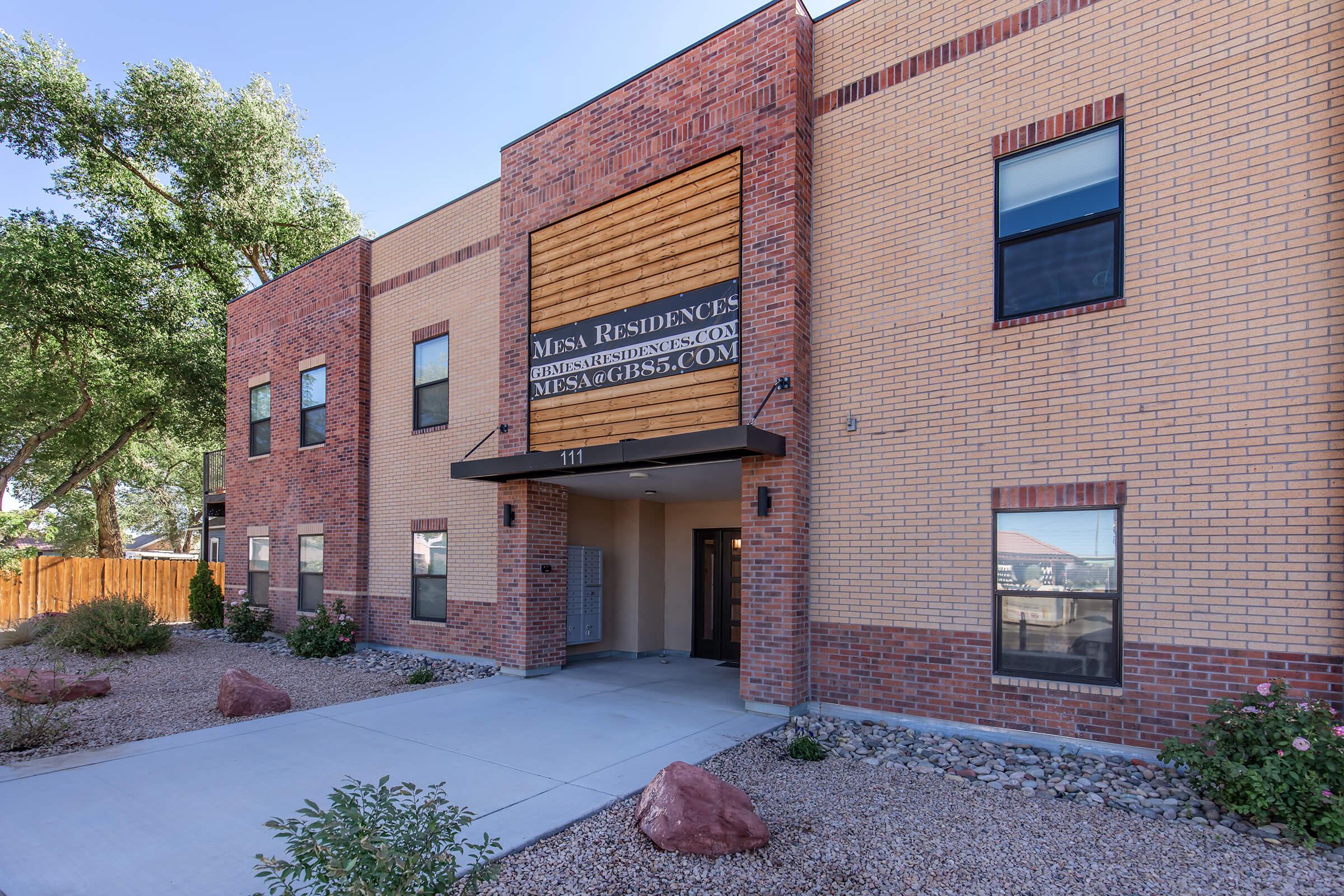 Exterior view of a modern brick building featuring a prominently displayed sign that reads "MESA RESIDENCE." The entrance is framed by a covered porch, with landscaping visible on either side, and a clear blue sky above. The building has a contemporary design with large windows.