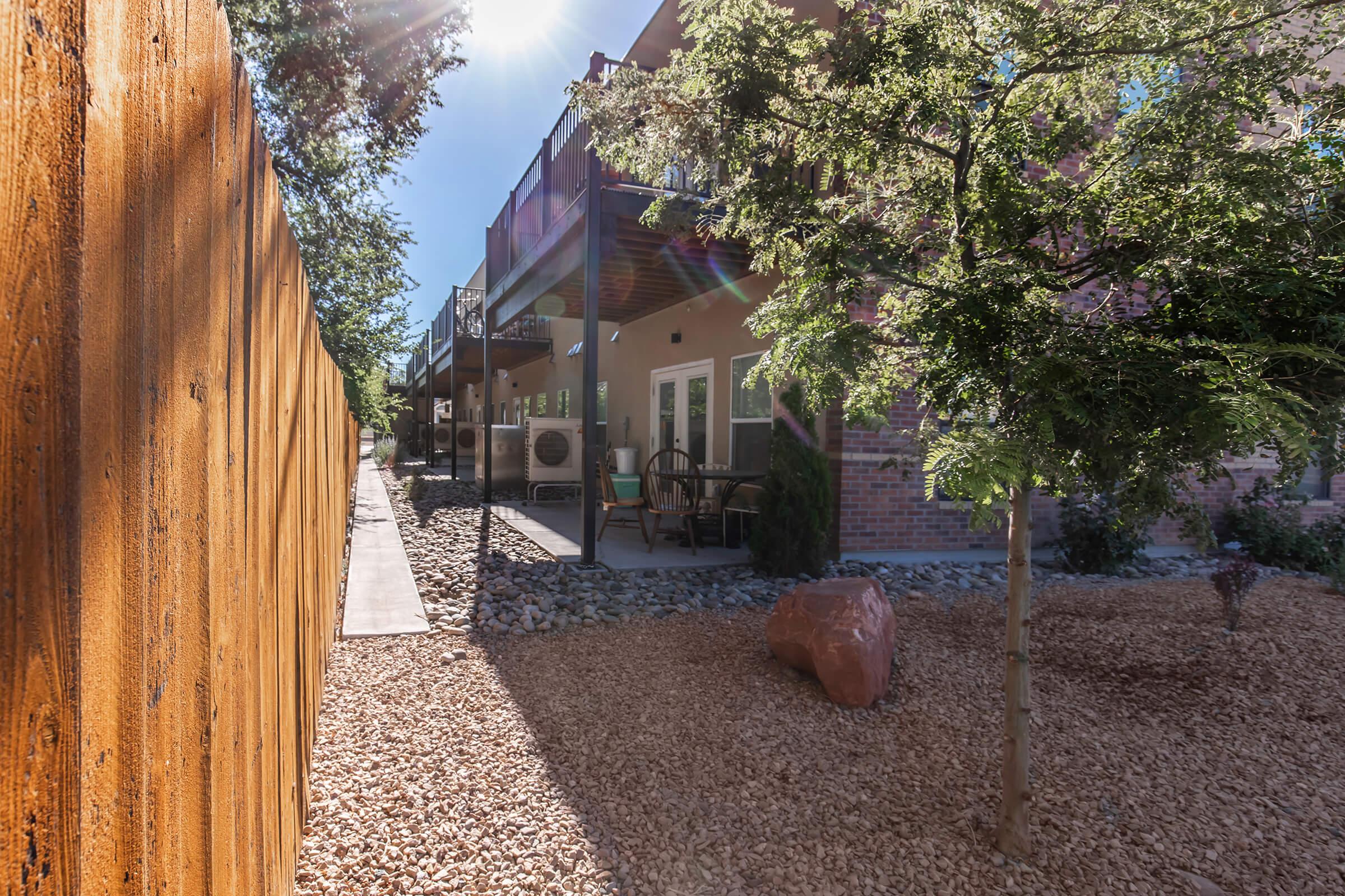 A scenic view of a landscaped area beside a building, featuring a wooden fence on the left, a pathway leading to the entrance, and a small rocky garden with a few trees. Sunlight shines brightly, creating a warm, inviting atmosphere.