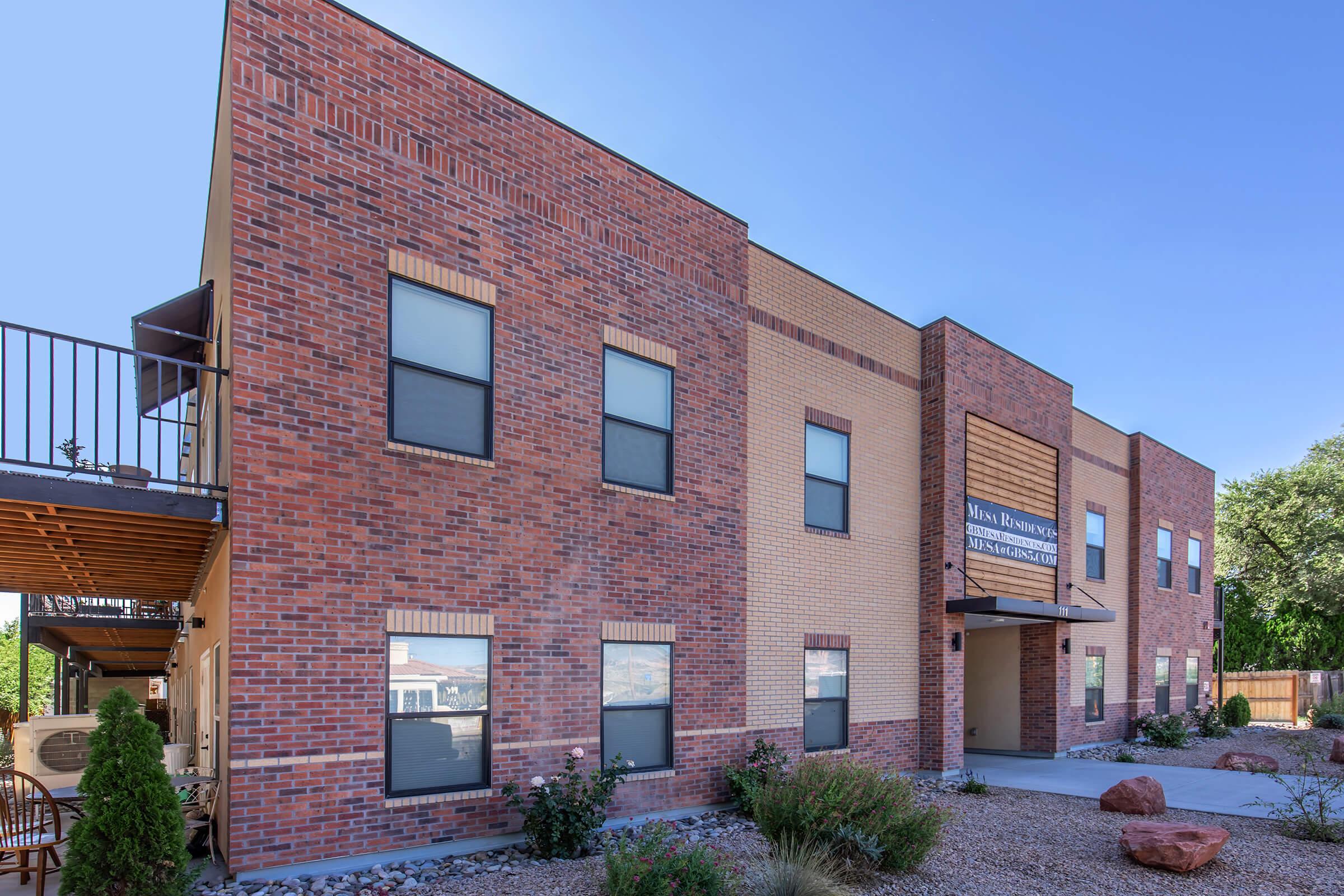A modern brick building with large windows and a welcoming entrance. The structure has light and dark brick sections, surrounded by landscaped gravel and rocks. The sky is clear and blue, indicating a sunny day.