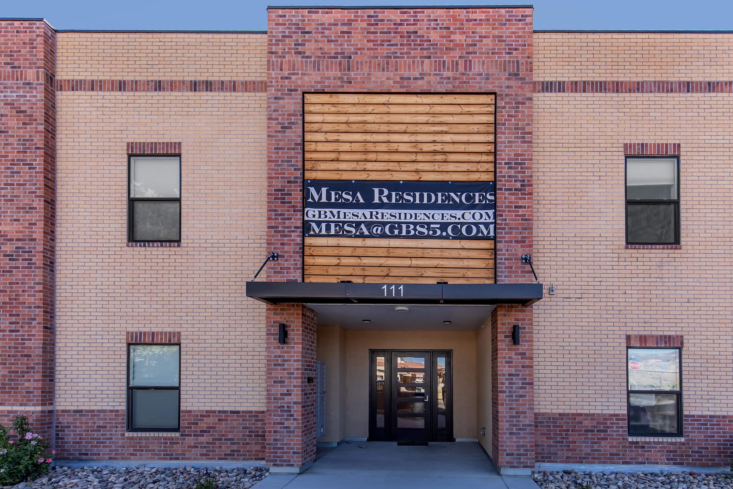 Entrance of Mesa Residences building featuring a wooden facade and a large sign displaying the website and email contact information. The structure has a brick exterior, with multiple windows on either side and a small overhang above the entrance door. The address displayed is 111.