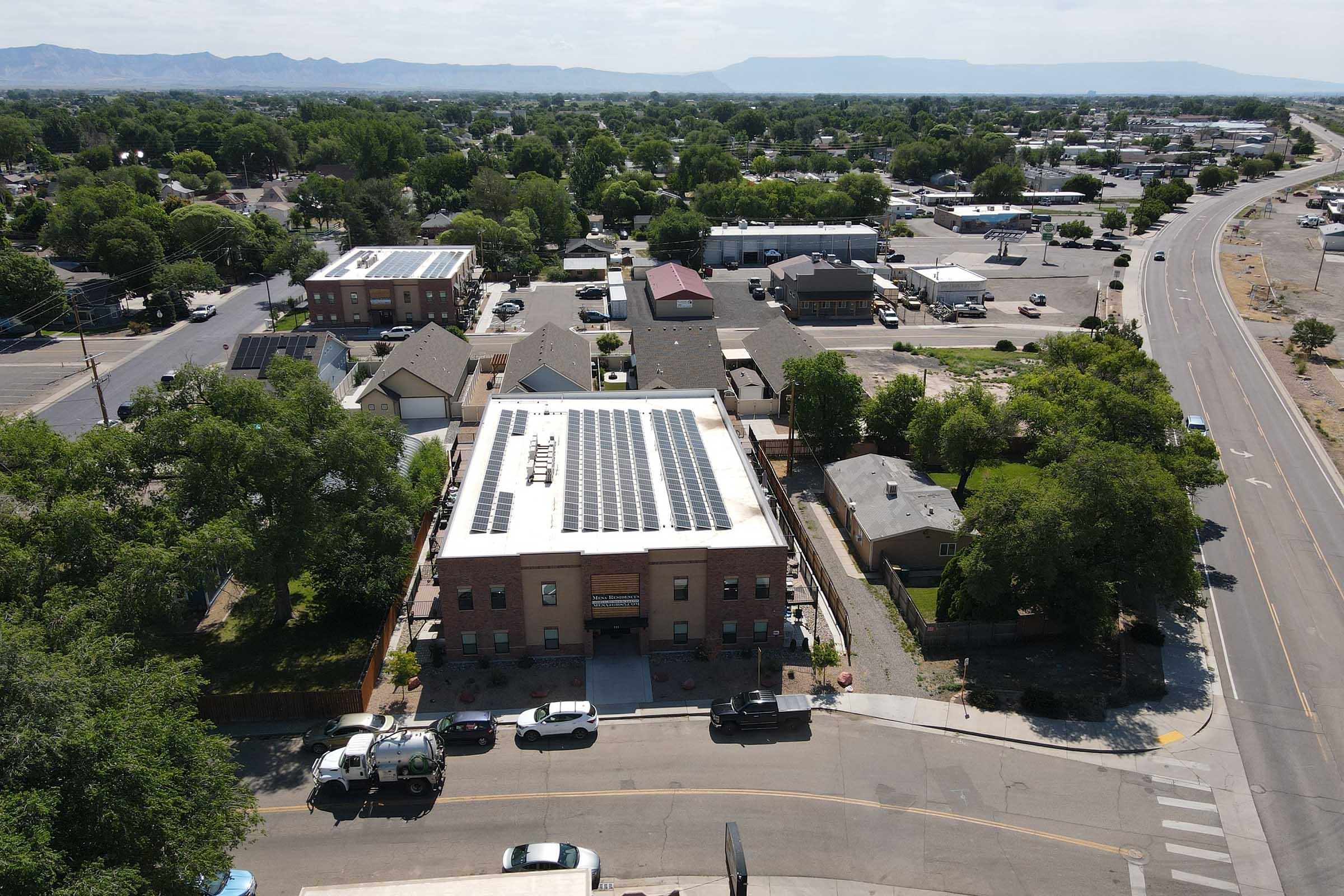 Aerial view of a neighborhood featuring a brick building with solar panels on its roof. Surrounding homes and a tree-lined street can be seen, along with sparse traffic. In the background, distant mountains are visible under a clear sky.