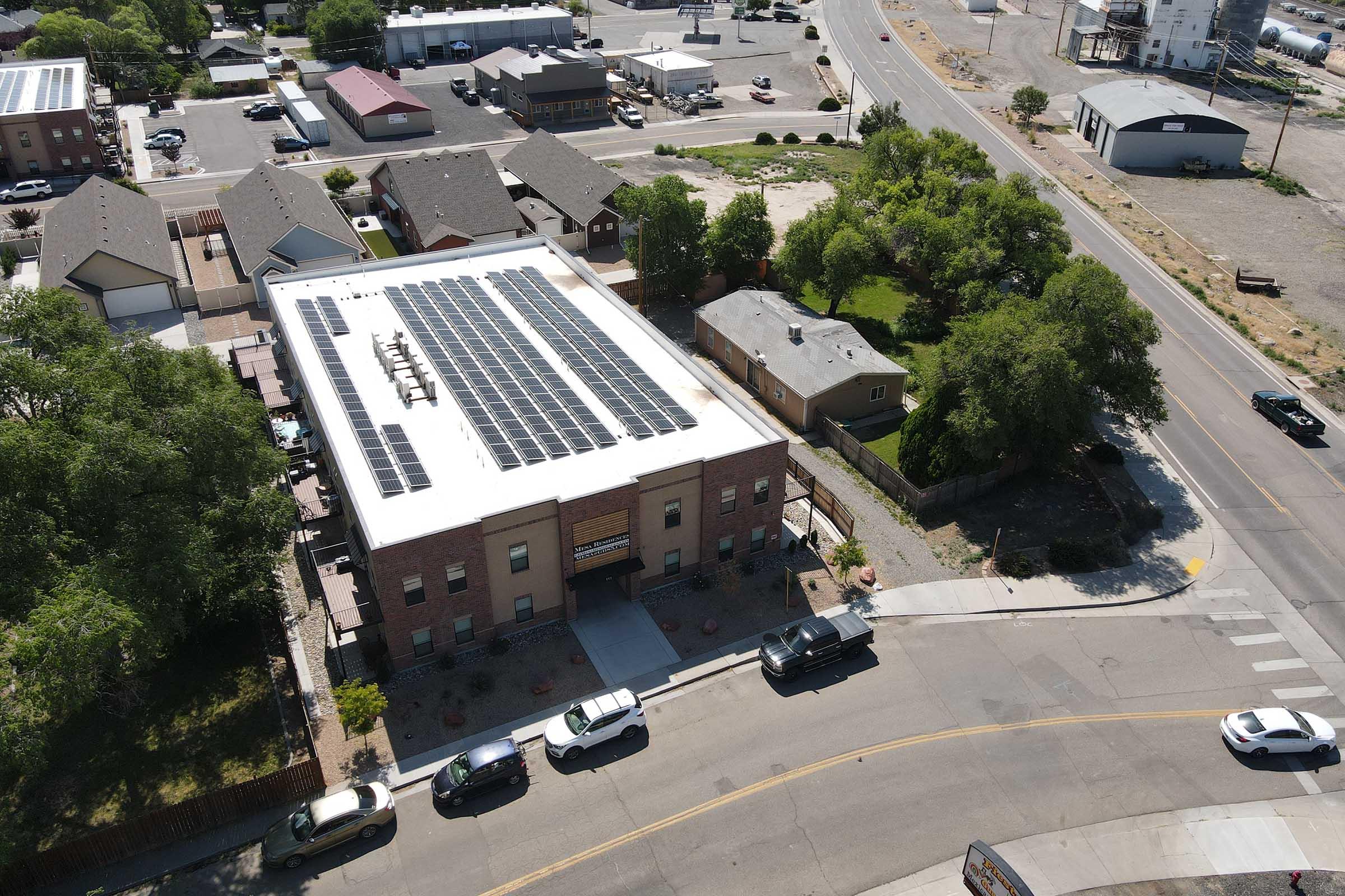 Aerial view of a building with solar panels on its roof, surrounded by residential and commercial properties. The street shows parked cars and a clear sky. Nearby trees and a road are visible, indicating a suburban setting.