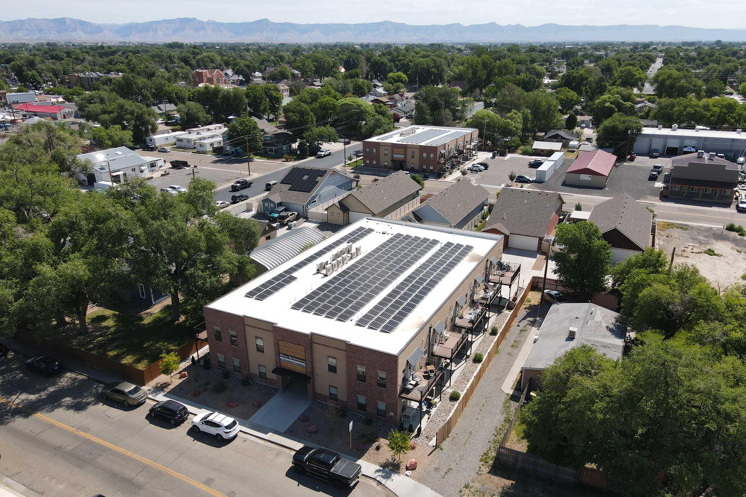 Aerial view of a building with a solar panel-covered roof, surrounded by residential and commercial properties, with mountains in the background. The scene captures a sunny day, showcasing a mix of architecture and greenery in the neighborhood.