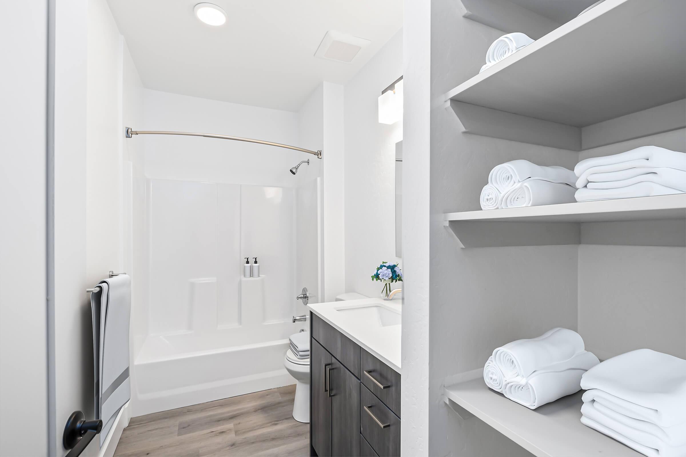 A clean, modern bathroom featuring a shower-tub combo, a white countertop sink with a mirror above, and neatly arranged white towels on open shelves. The floor is wooden, and there's a small decorative element, like flowers, on the countertop, suggesting a fresh and inviting space.