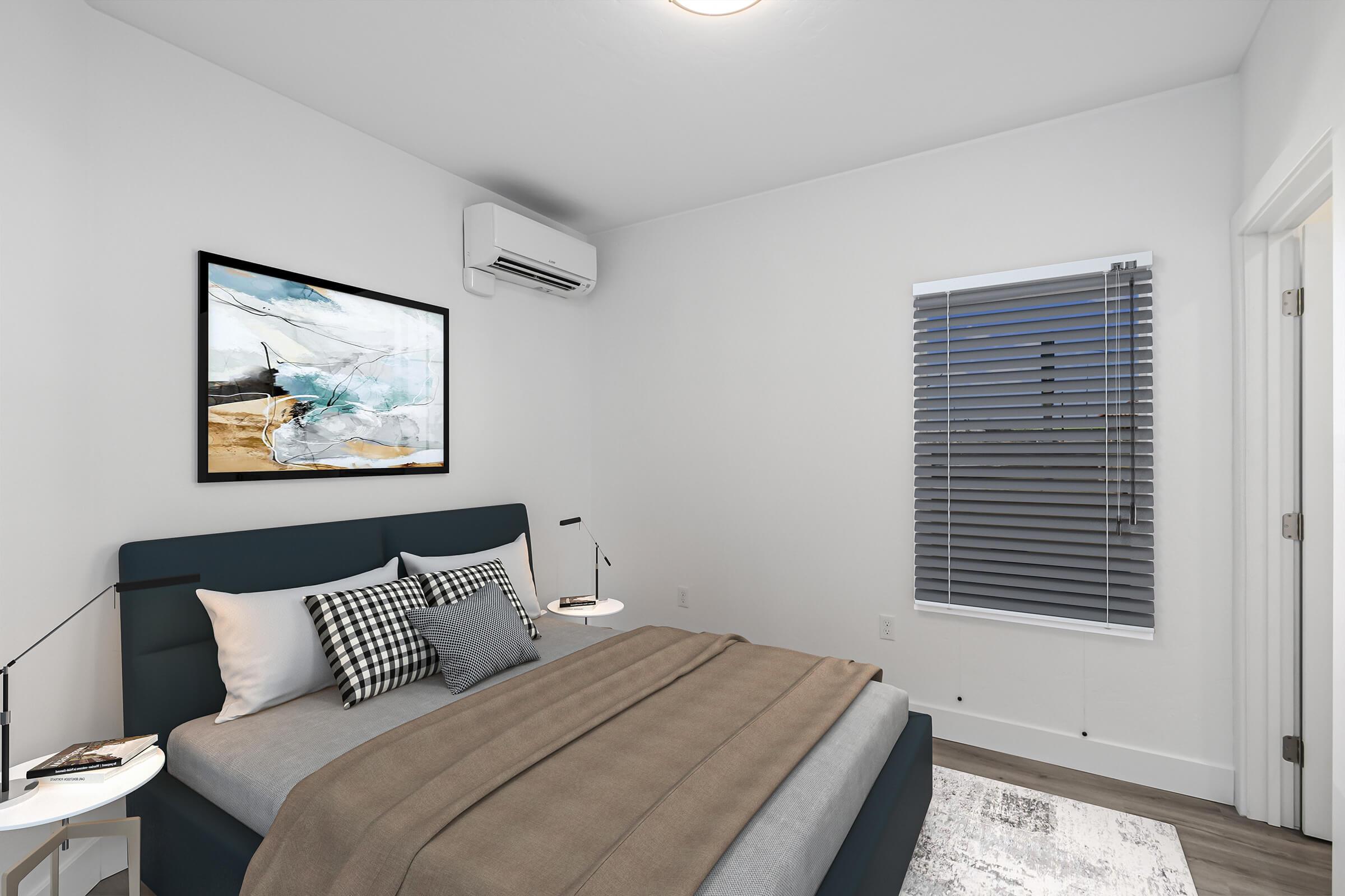 A modern bedroom featuring a dark blue bed with gray bedding and decorative pillows, a sleek nightstand with a lamp, and a piece of abstract art on the wall. The room has a large window with blinds, an air conditioning unit on the wall, and light-colored walls, creating a cozy atmosphere.