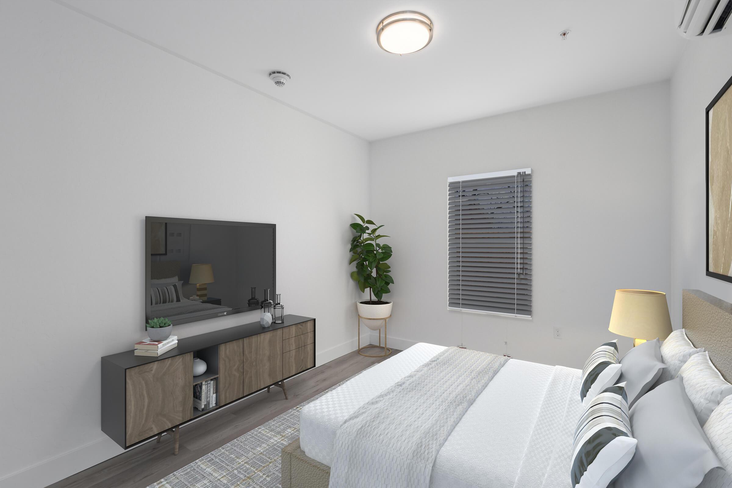 A modern bedroom featuring a neatly made bed with light-colored linens, a stylish bedside lamp, and a large wall-mounted television on a sleek media console. A potted plant adds a touch of greenery, while a window with blinds allows natural light to enter, enhancing the serene atmosphere.