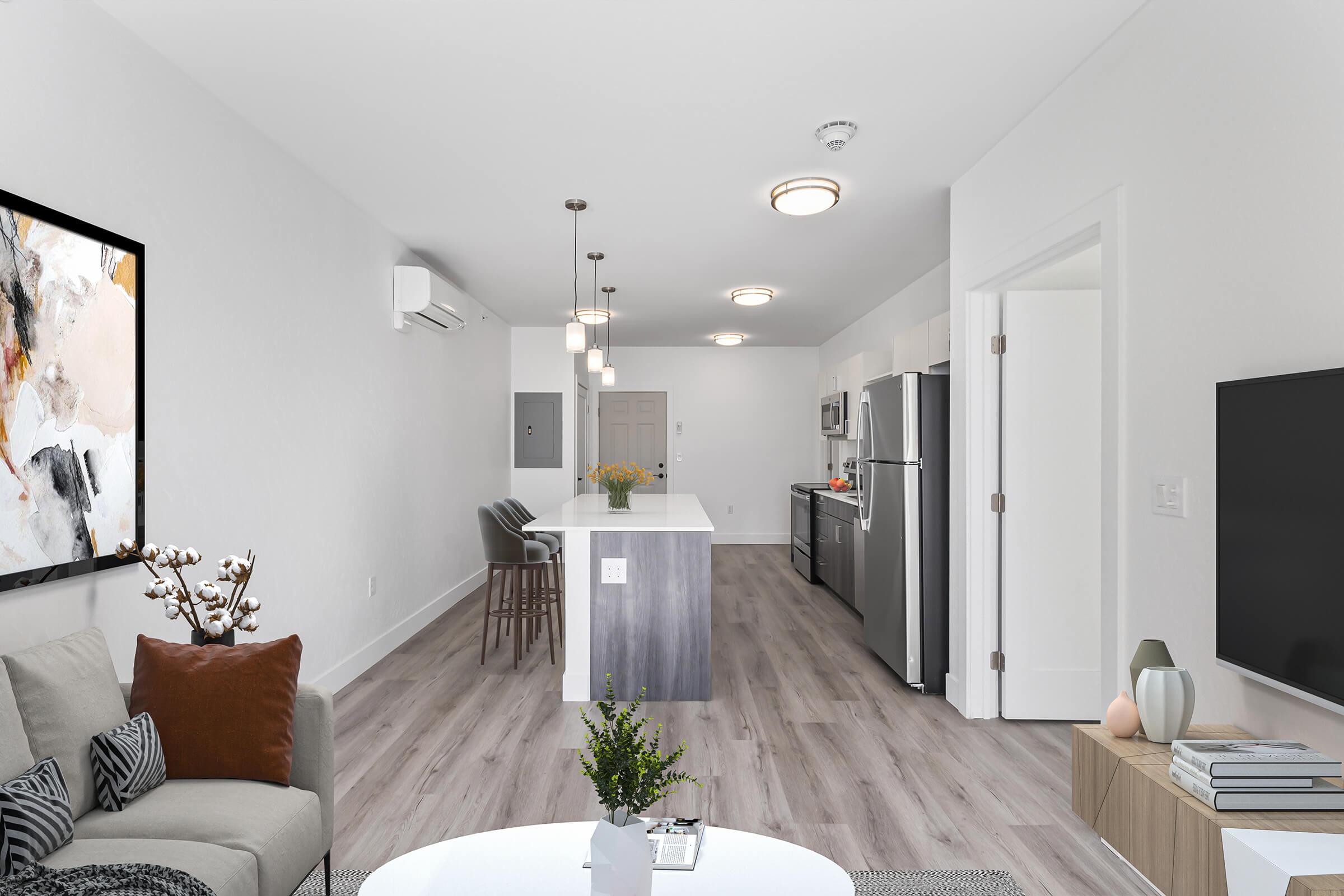 A modern, open-concept living space featuring a cozy sofa with decorative pillows, a coffee table, and a dining area with bar stools. Bright overhead lights illuminate the room, which includes a kitchen with stainless steel appliances, a central island, and large windows allowing natural light to enter.