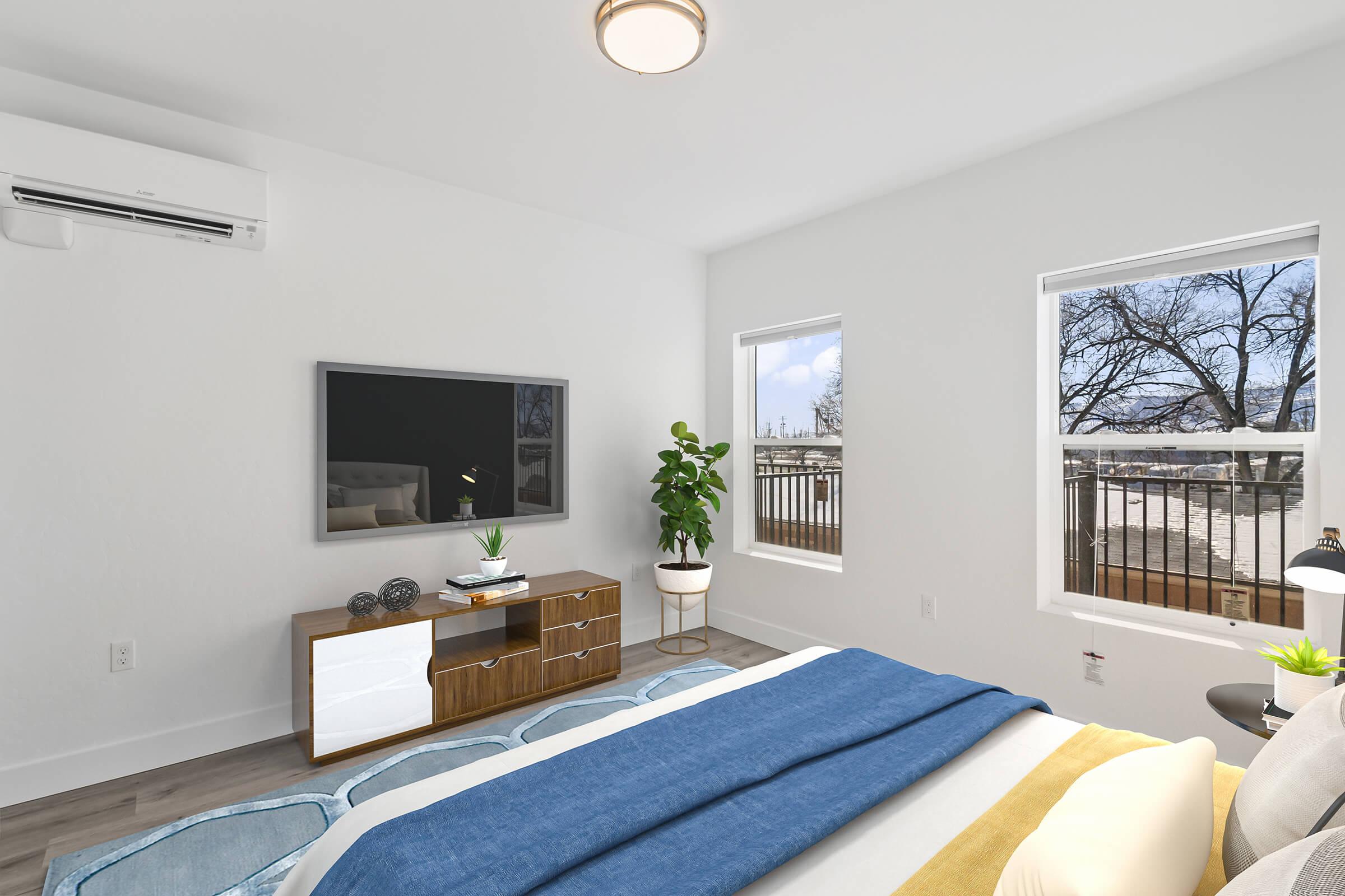 A modern bedroom featuring a bed with a blue throw blanket and yellow pillows. There is a stylish wooden dresser with a TV mounted above it. A potted plant adds a touch of greenery. Large windows provide natural light and a view of the outdoors, creating a bright and inviting atmosphere.