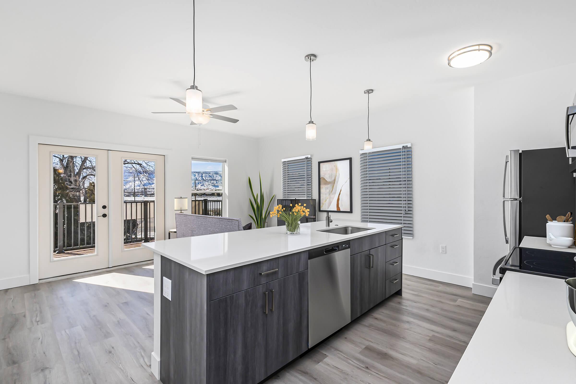 Modern kitchen featuring a large island with a sink and dishwasher, stainless steel appliances, and pendant lighting. Large windows provide natural light, with a view of a balcony and outdoor space. Light wood flooring complements the contemporary design, and a vase of yellow flowers adds a touch of color.