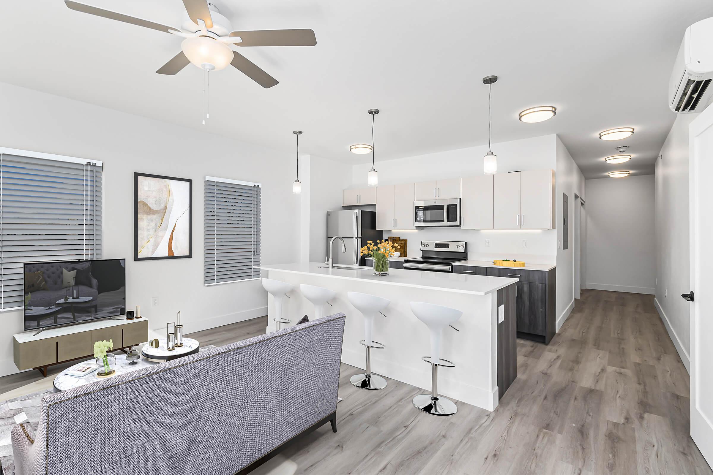 A contemporary kitchen and living area featuring a stylish gray sofa, a flat-screen TV on a light wood console, and a modern kitchen bar with three white stools. The kitchen includes stainless steel appliances, white cabinetry, and pendant lighting. Large windows allow natural light to fill the space.