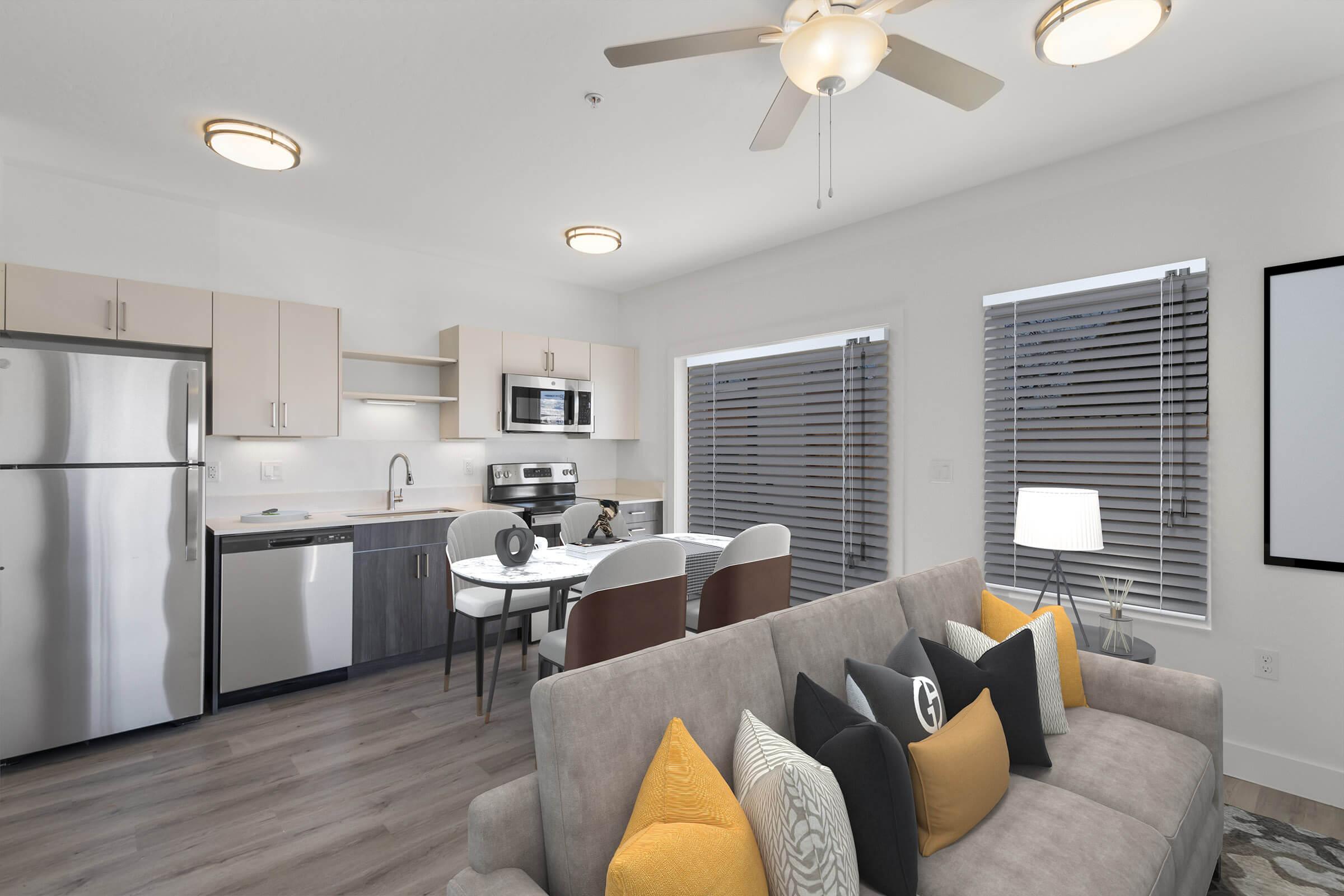 A modern living area featuring a gray sofa adorned with colorful cushions. The open kitchen includes stainless steel appliances, a dining table, and large windows with blinds. The space is well-lit with ceiling lights, offering a clean and contemporary aesthetic.