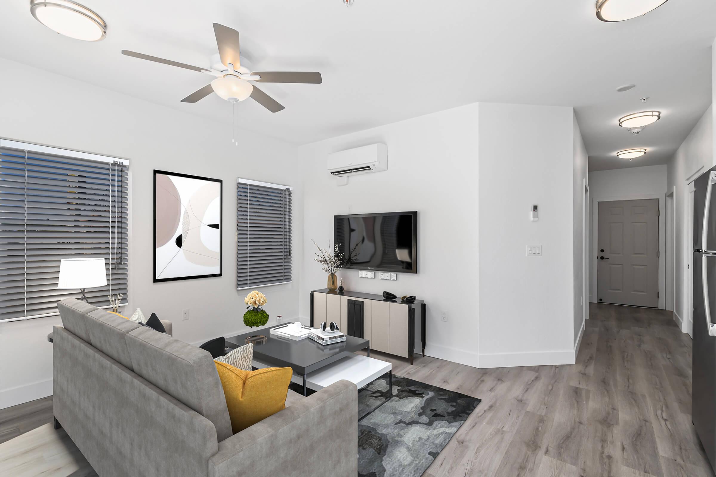 Modern living room featuring a light gray sofa with yellow accents, a coffee table, and a wall-mounted TV. The space has large windows with blinds, a ceiling fan, and contemporary art on the wall. A small hallway is visible in the background, leading to a door. The flooring is a light wood laminate.