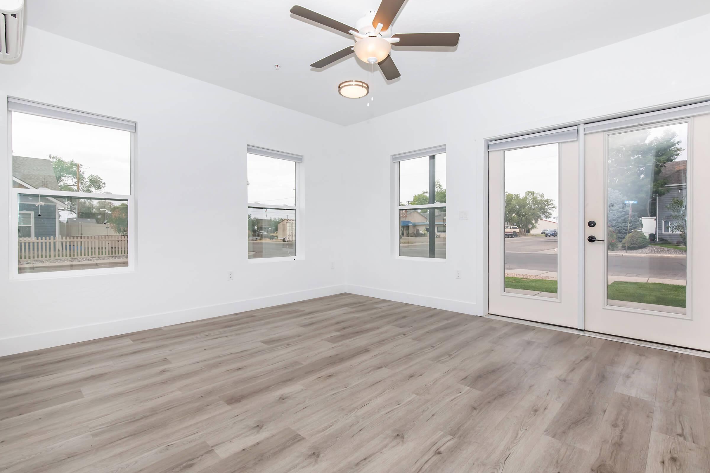 A bright, empty living room featuring light gray flooring, white walls, and a ceiling fan. There are two windows on one side and a pair of sliding glass doors leading outside, allowing natural light to flood the space. The overall atmosphere is open and inviting, perfect for personal decor and furniture.